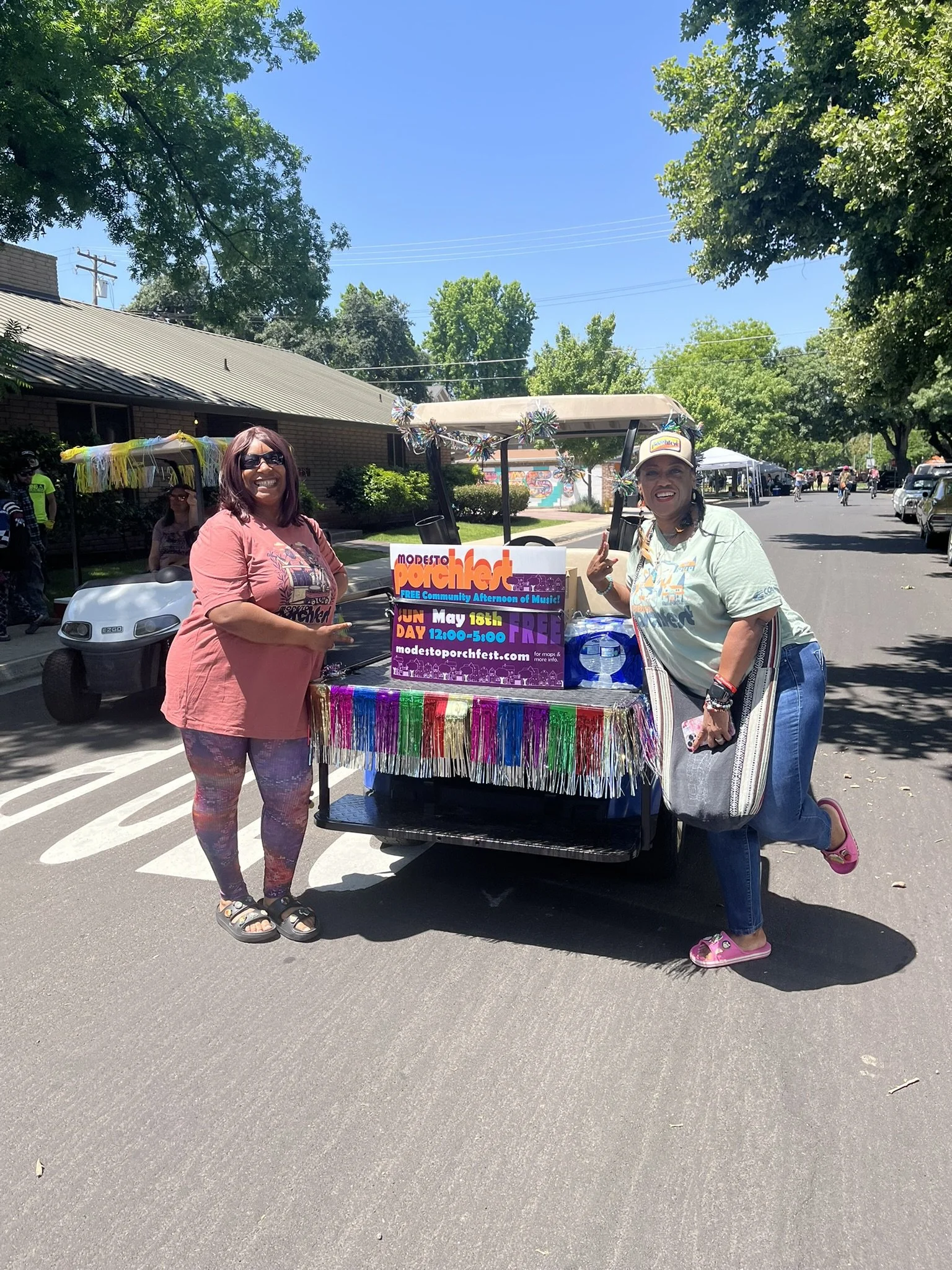 Two women standing beside a colorful cart at a community festival, with trees and tents visible in the background.