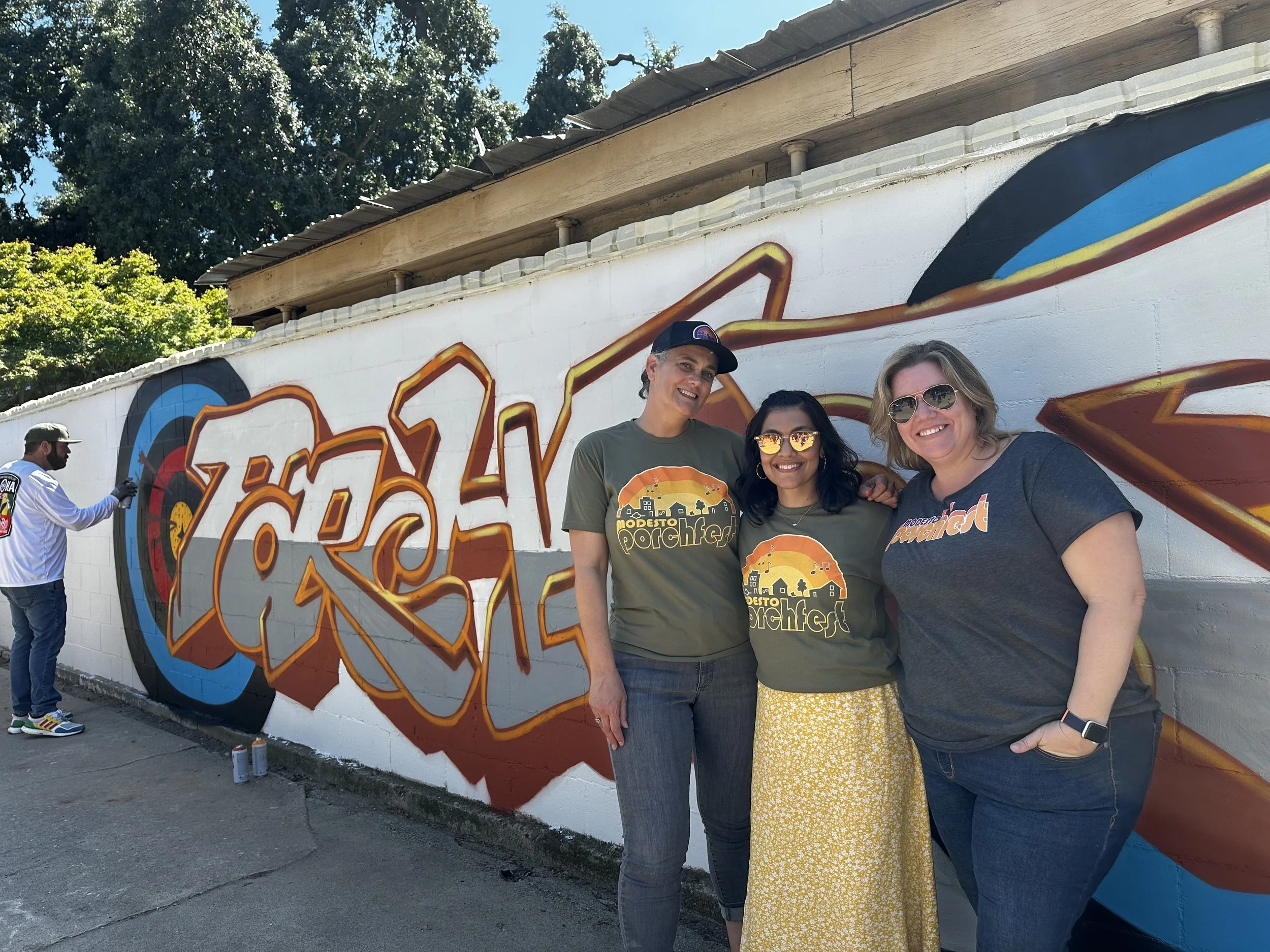 Three women stand in front of a mural with graffiti-style artwork, smiling for the photo. Another man is seen on the left spray painting the wall with graffiti. The women are wearing matching gray and yellow T-shirts promoting Modesto Porchfest, with one in sunglasses and a yellow skirt.