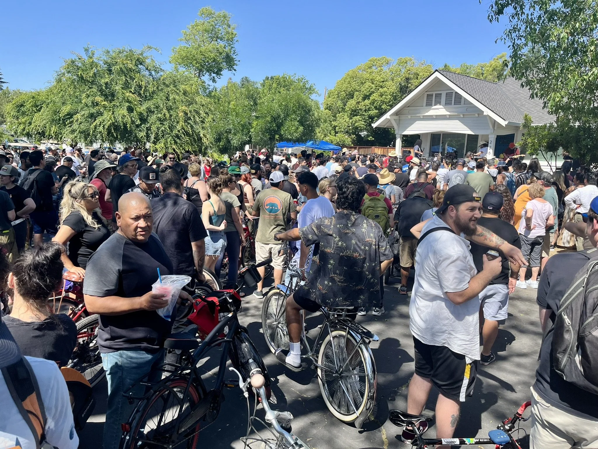 Large crowd of people gathered outdoors in front of a house, some holding drinks, others standing or sitting with bicycles, during a sunny day with blue sky and green trees.