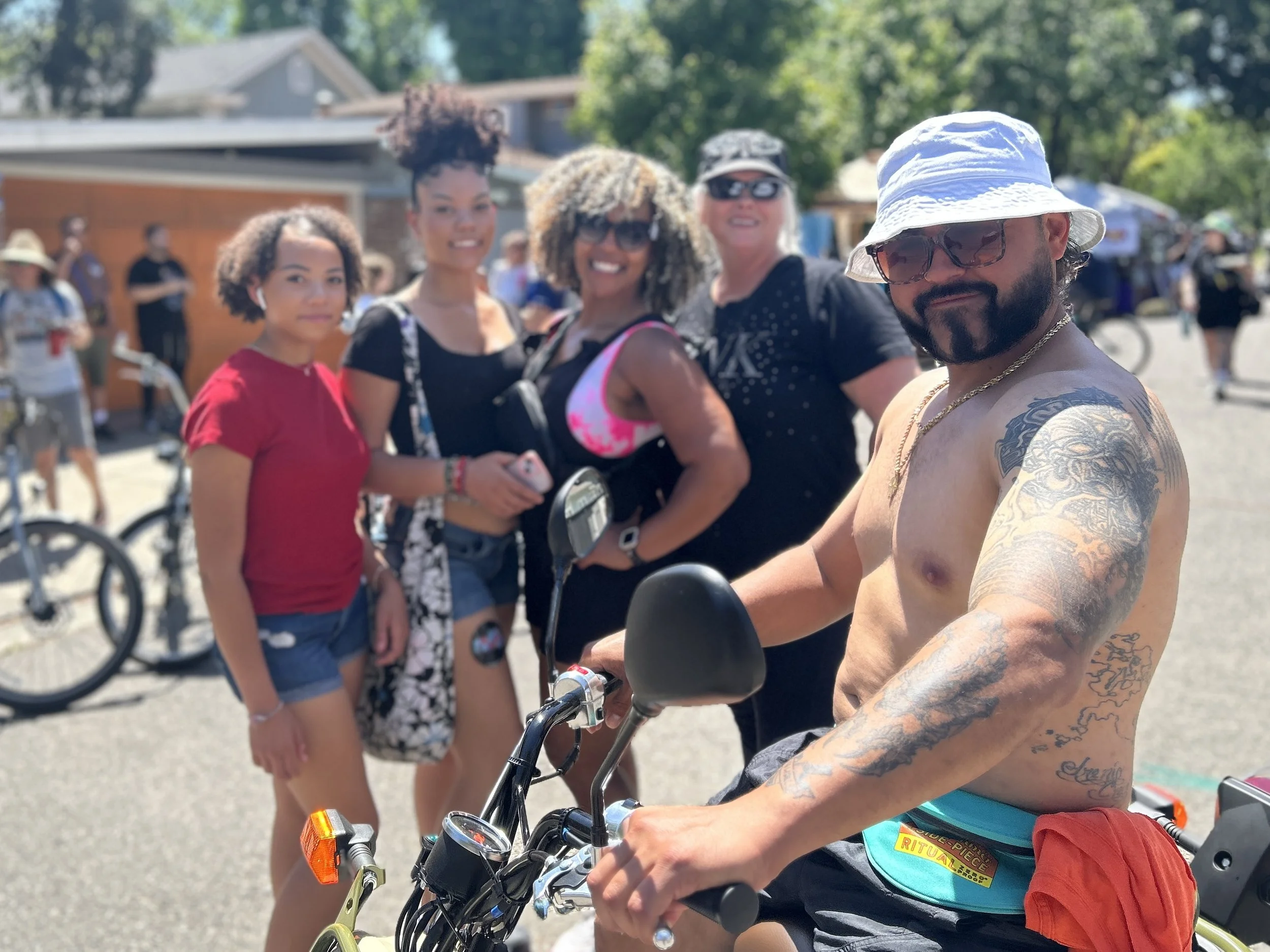 Group of five diverse people standing outdoors on a sunny day, with some holding bicycles at an outdoor event or market.