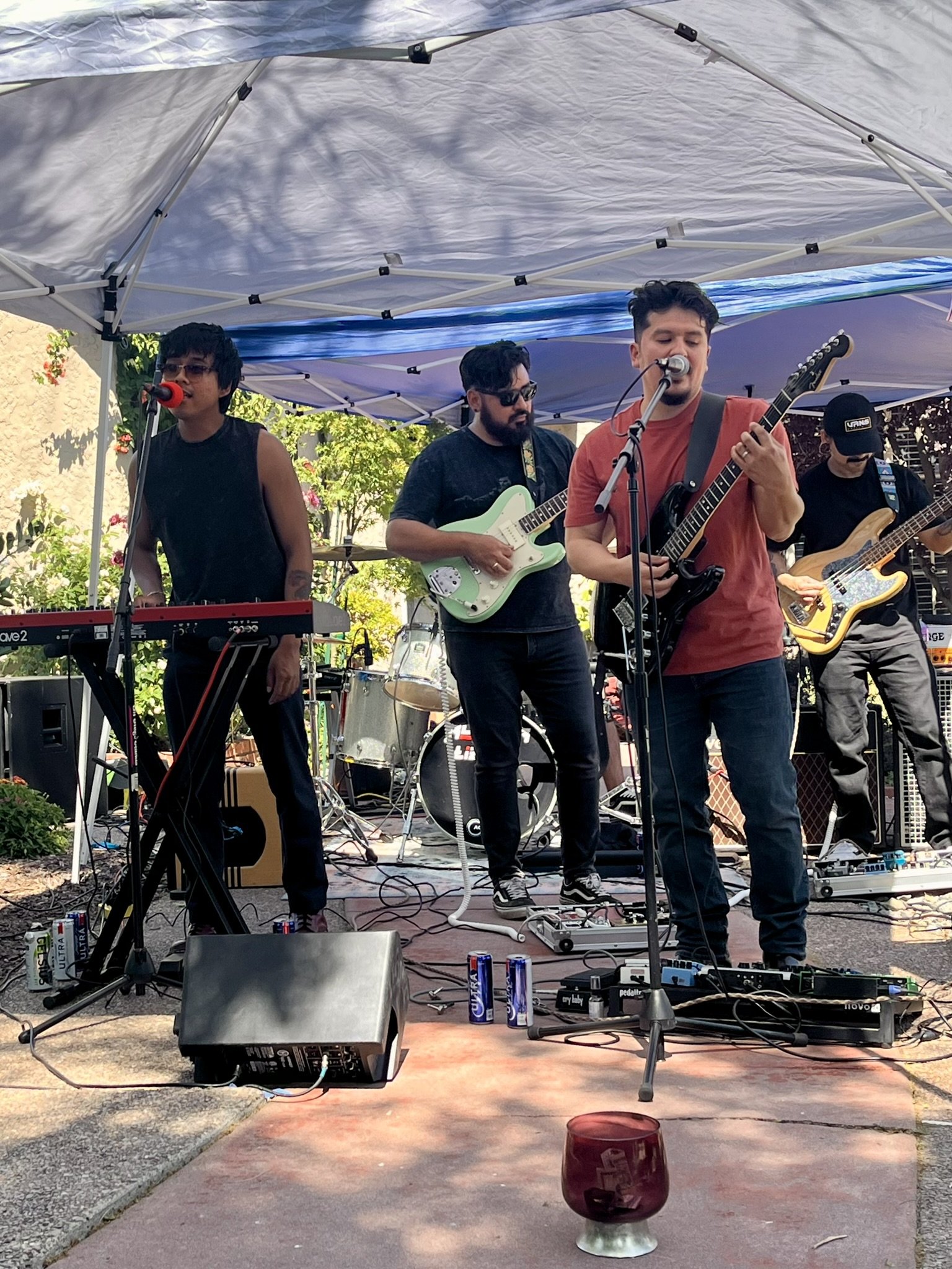 Four musicians perform under a tent outdoors, with a keyboard, electric guitars, and a bass. There are drinks on the ground, and trees and flowers are in the background.