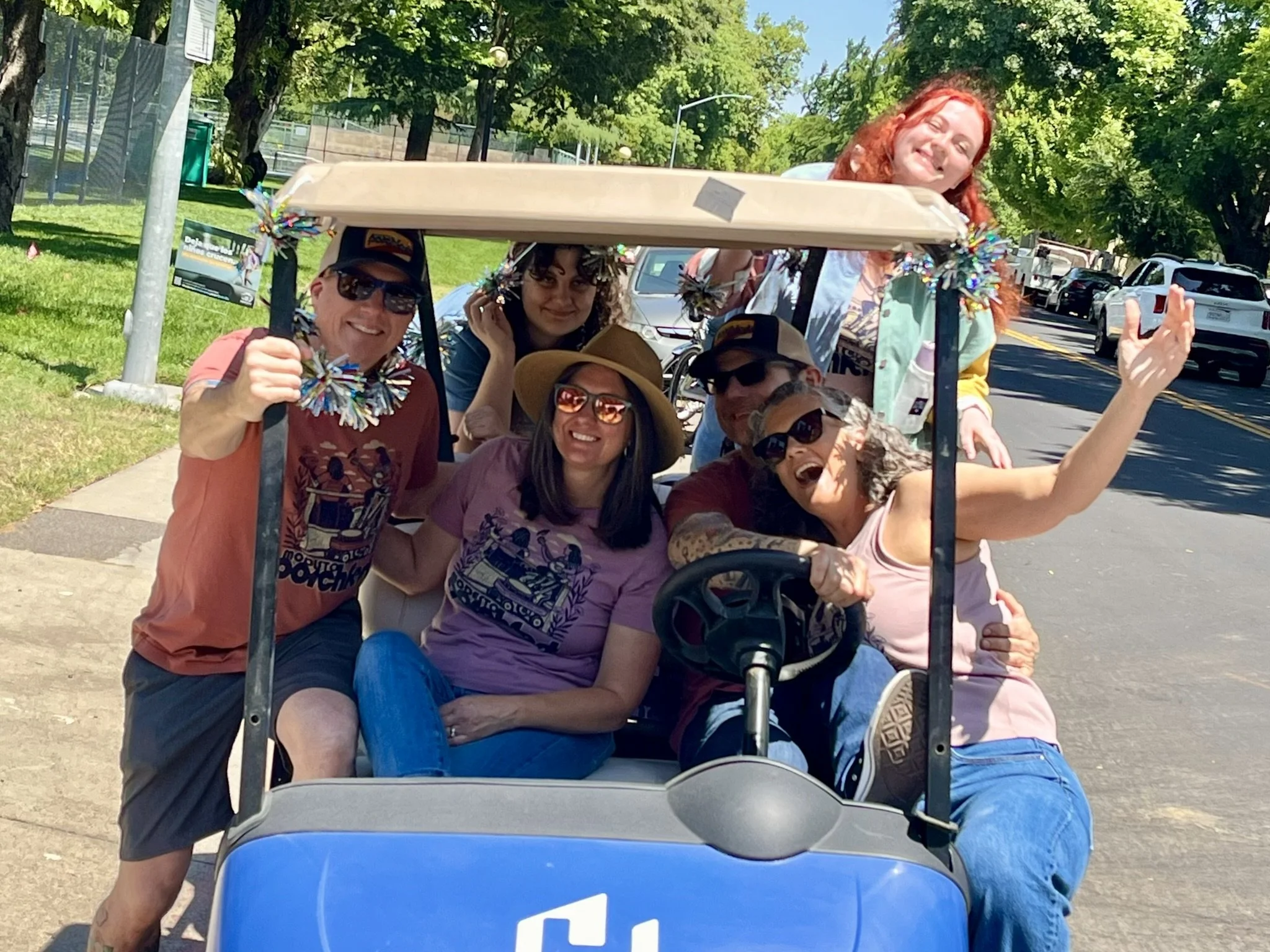 Group of six people smiling and posing on a golf cart with a canopy decorated with colorful tinsel on a sunny day. They are enjoying an outdoor event, with green trees and parked cars visible in the background.
