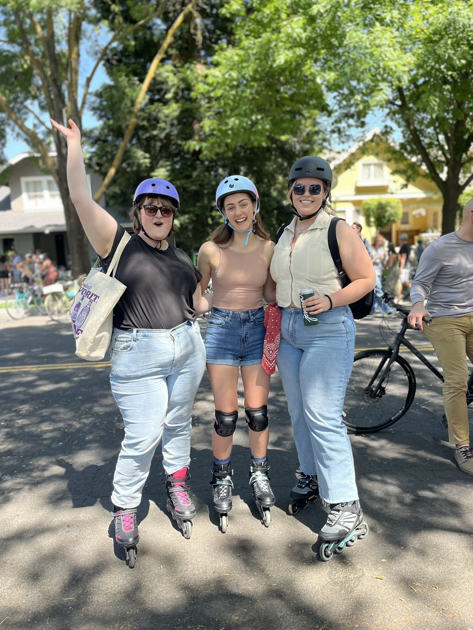 Three women wearing helmets rollerblading on a street, smiling and posing for the photo, with trees and houses in the background, sunny day.