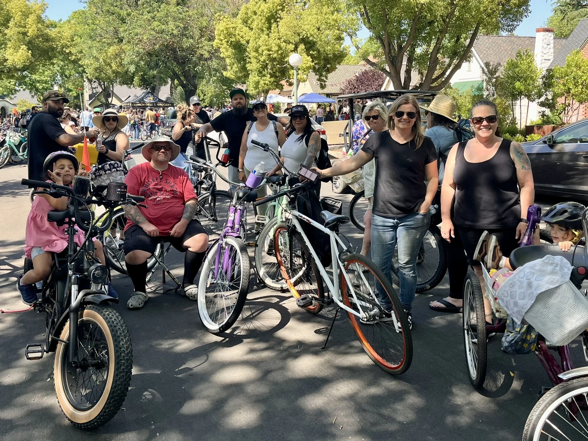 A group of people gathered outdoors with bikes, some smiling, in a sunny park with trees and houses in the background.