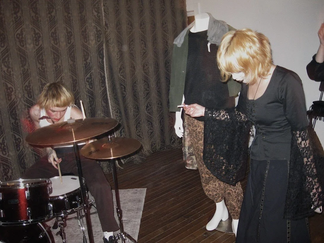 Man with blond hair playing drums and a girl dressed in all black with the same hair colour and style is smoking a cigarette nearby.
