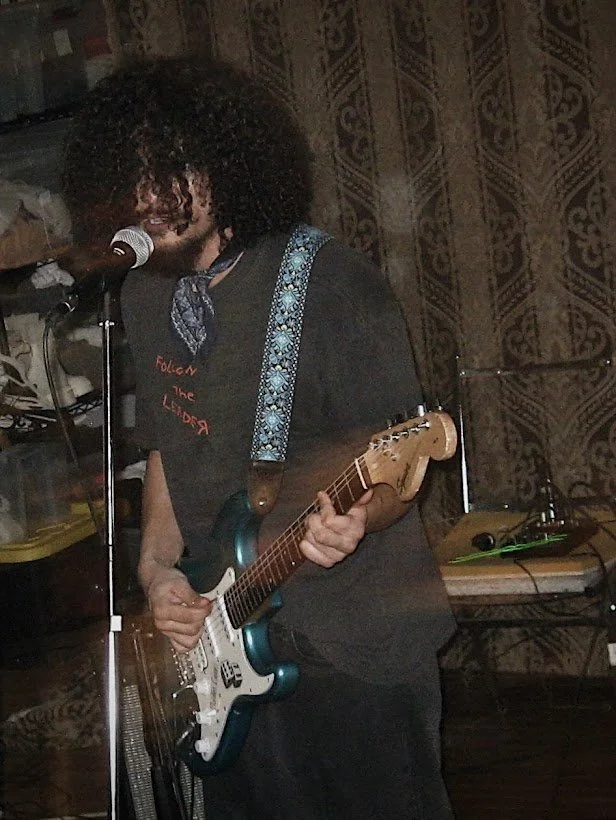 Man with curly hair singing into a microphone and playing electric guitar on stage at a concert. Shot in black and white.