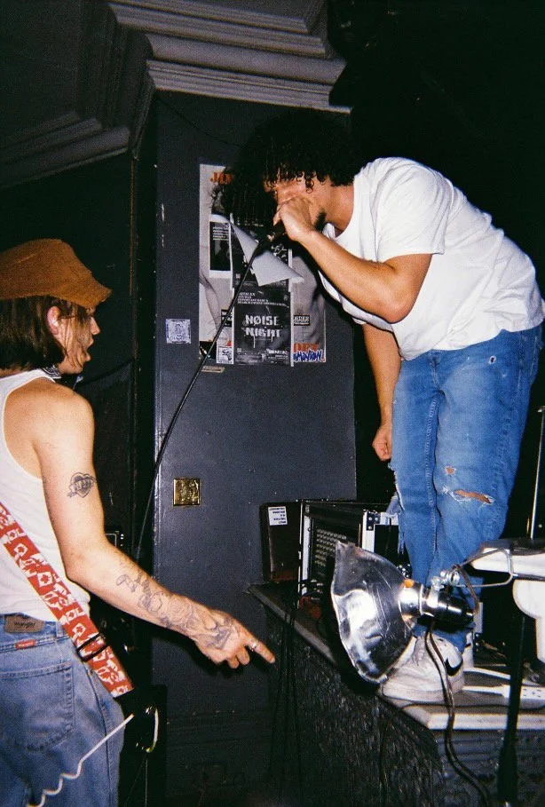 Two young men performing music in a small venue. One is standing on a table, singing into a microphone, while the other is pointing and looking at him. The environment has posters on the wall, and music equipment is visible.