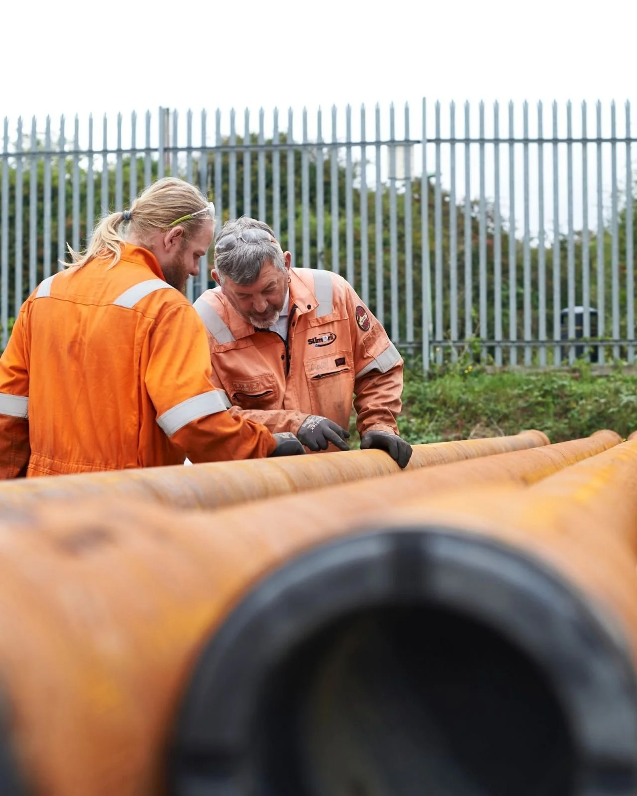 Two workers in orange safety uniforms inspect a large pipe outdoors near a tall metal fence and greenery.