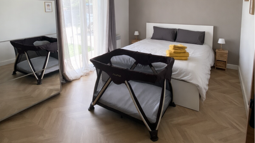 Chambre avec lit double, deux tables de nuit avec lampe, deux serviettes jaunes pliées sur le lit, nacelle de bébé devant le lit, armoire avec miroir.