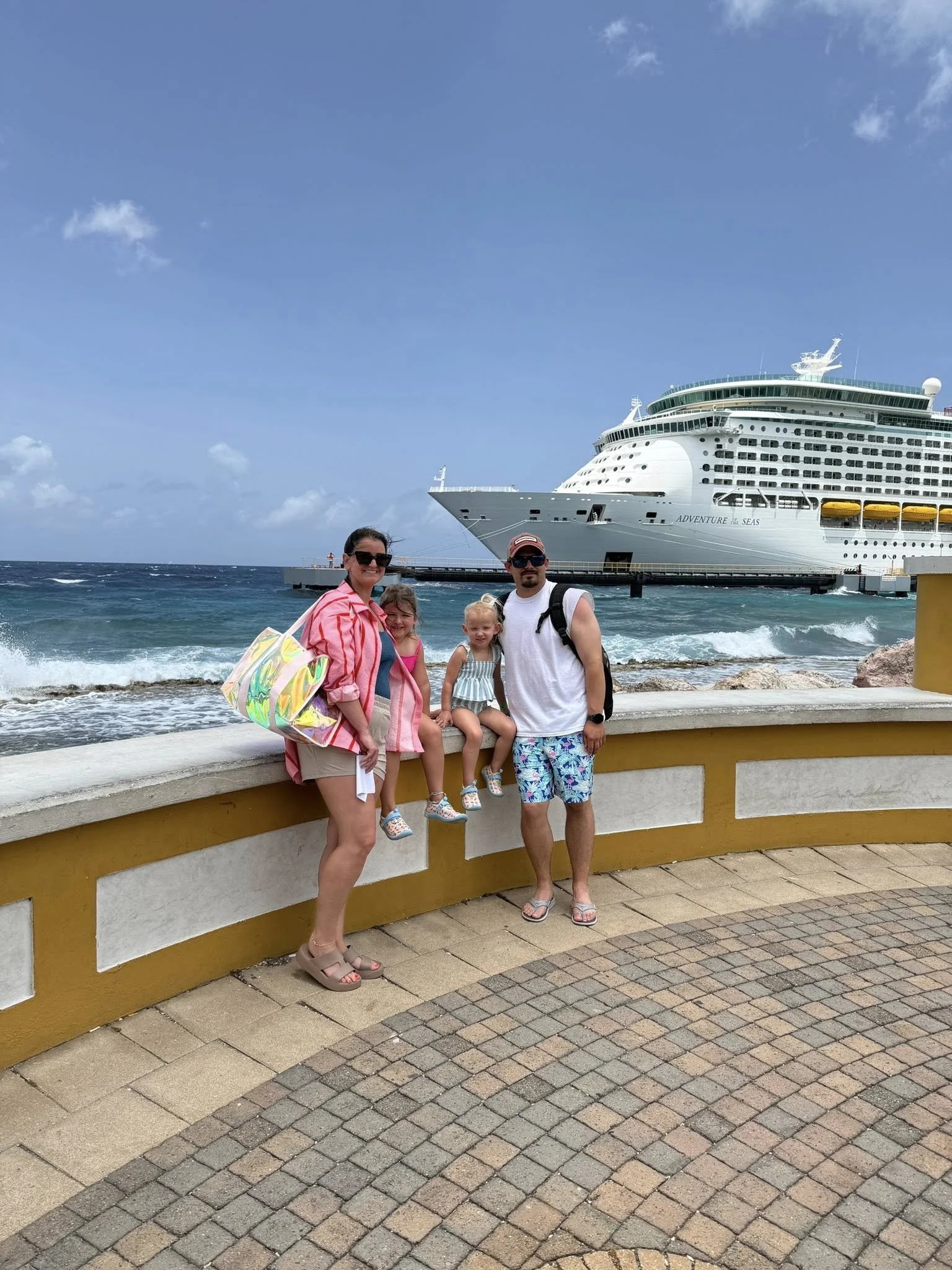 A family of four standing by a yellow and white wall on a pier, with a large cruise ship named 'Adventure of the Seas' in the background, and the ocean with waves and a partly cloudy sky.