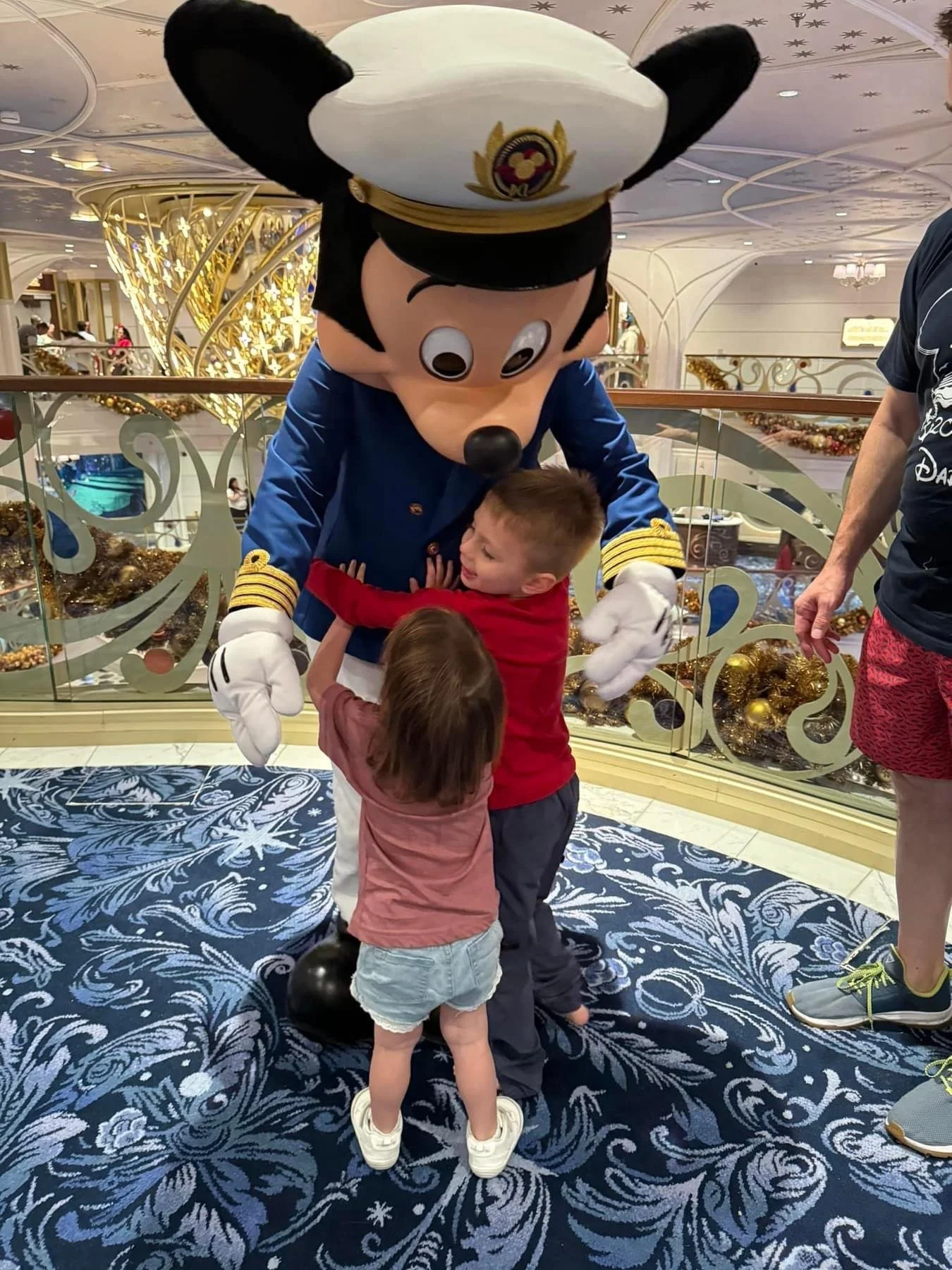 Two children hugging Mickey Mouse dressed as a ship captain on a Disney cruise decorated for the holidays.