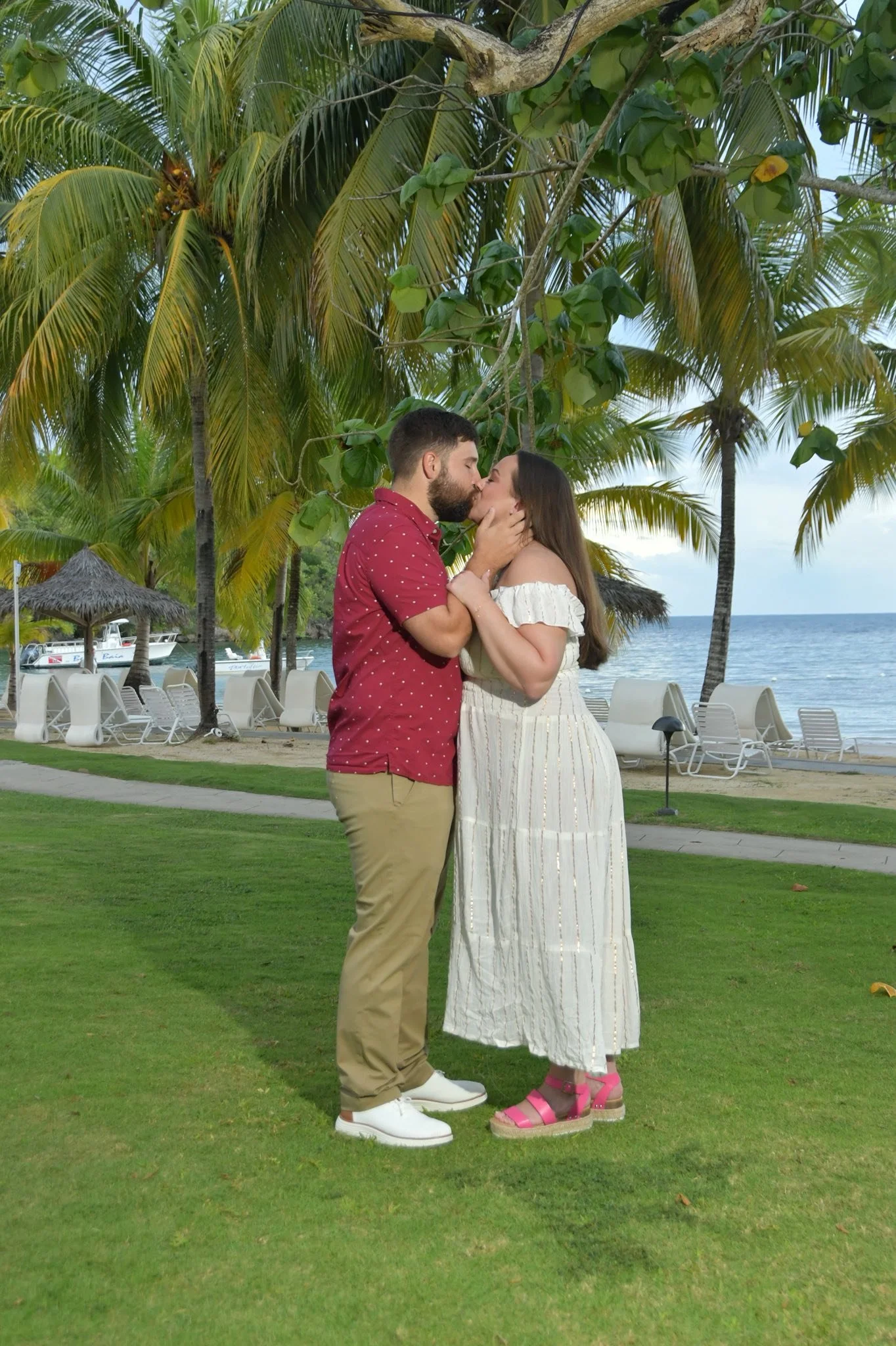 A honeymoon couple kissing on a lush, green lawn near a beach with palm trees and lounge chairs in Jamaica.
