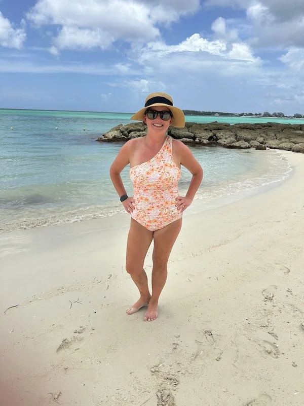 A woman in a floral swimsuit, wide-brimmed hat, and sunglasses standing on a sandy beach near rocks with the ocean and a partly cloudy sky in the background.