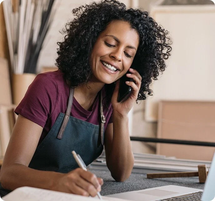 Woman smiling while talking on the phone and writing with a pen at a work desk.