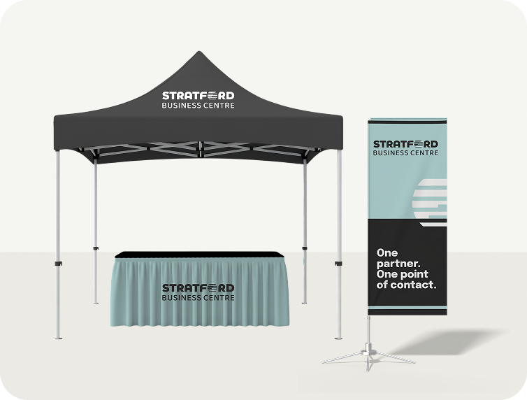 A white outdoor event tent with a table underneath, a blue vertical flag with an arrow and text, and a blue and black sign that says "Flag Mockup". 