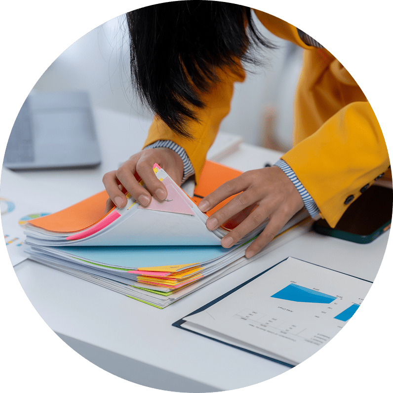 Person flipping through a large stack of colorful documents or notebooks at a desk, with papers and a laptop nearby.