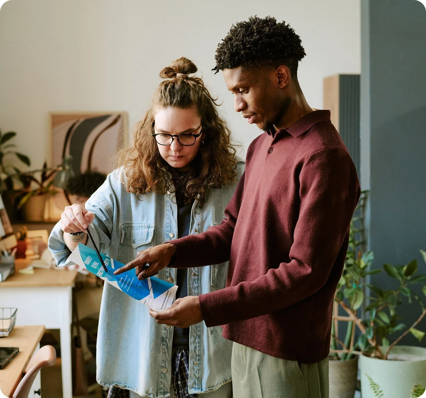 Two young adults, a woman with curly hair and glasses and a man with short curly hair, are examining a blue pamphlet or brochure together in an office or workspace setting.