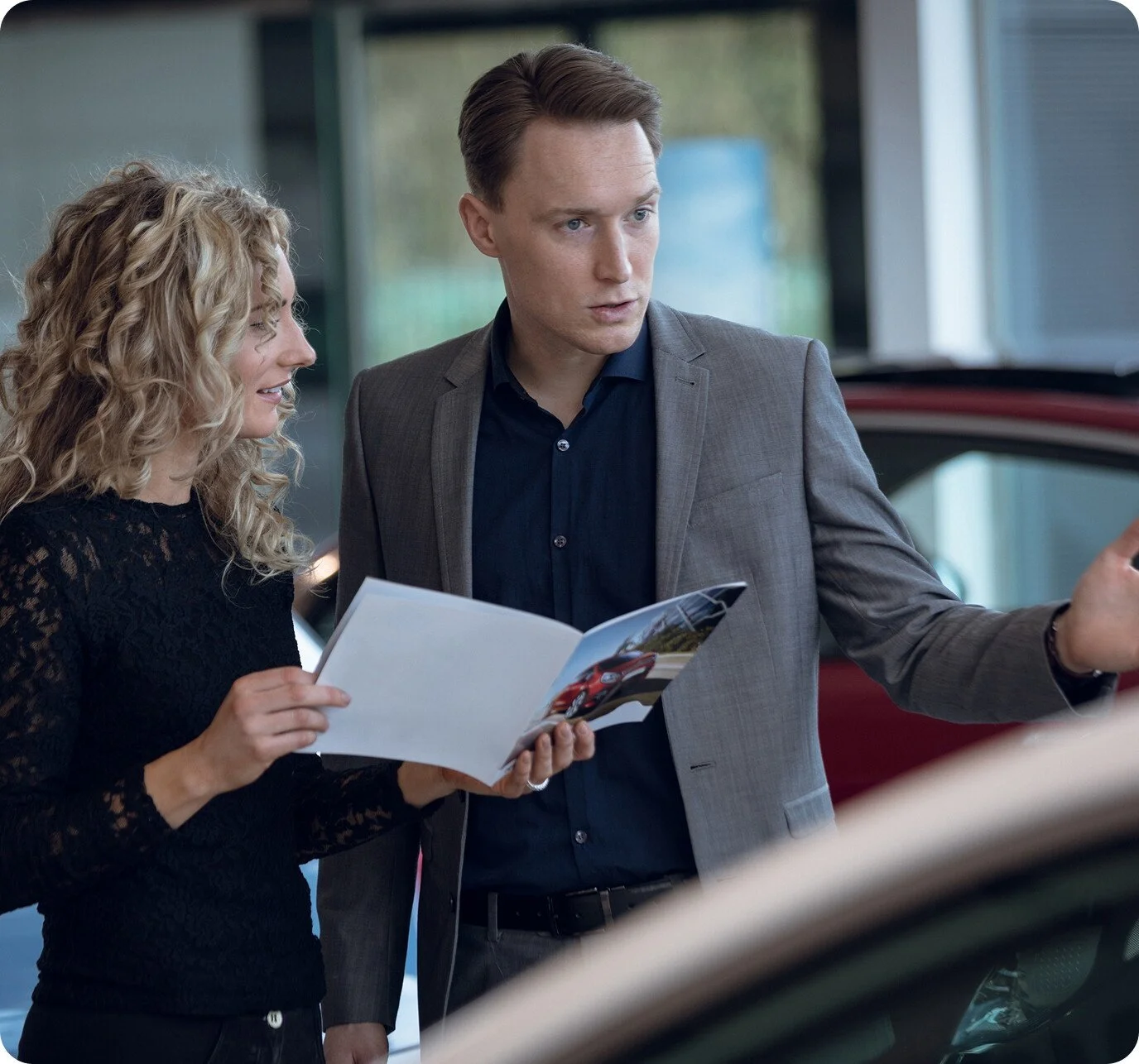 A man and woman standing next to a red car, looking at a brochure. The woman is holding the brochure and smiling, while the man is pointing at something outside the car.