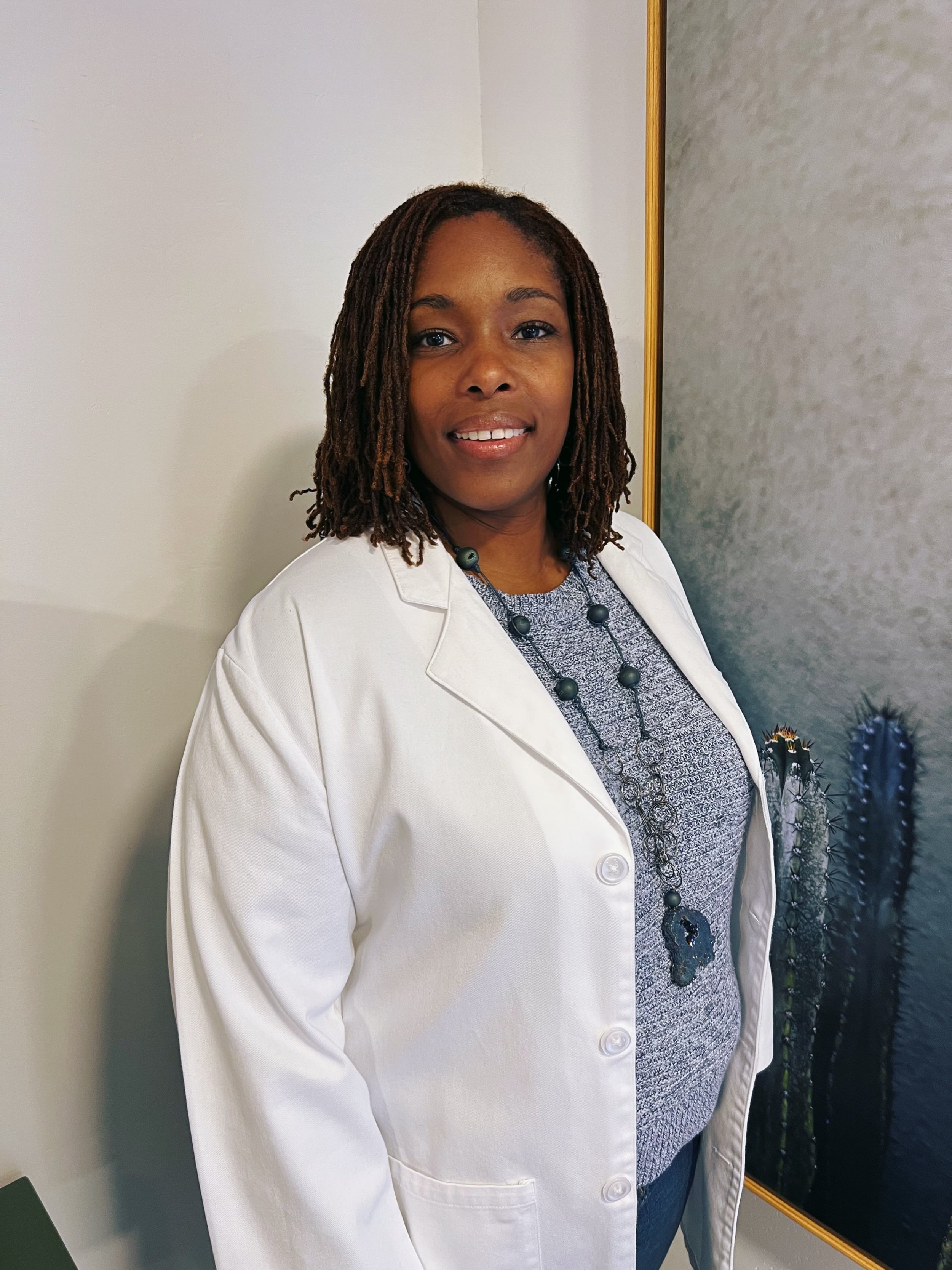 A woman with shoulder-length dreadlocks smiling, wearing a white blazer over a patterned top with a long necklace, standing in front of a wall with a framed artwork of cacti.