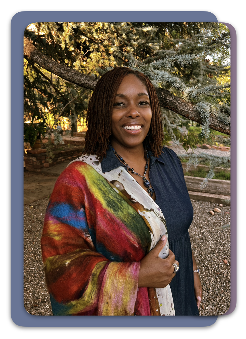 A woman with shoulder-length dreadlocks smiling outdoors, standing in front of a tree with dense green foliage, wearing a colorful scarf and a dark dress.