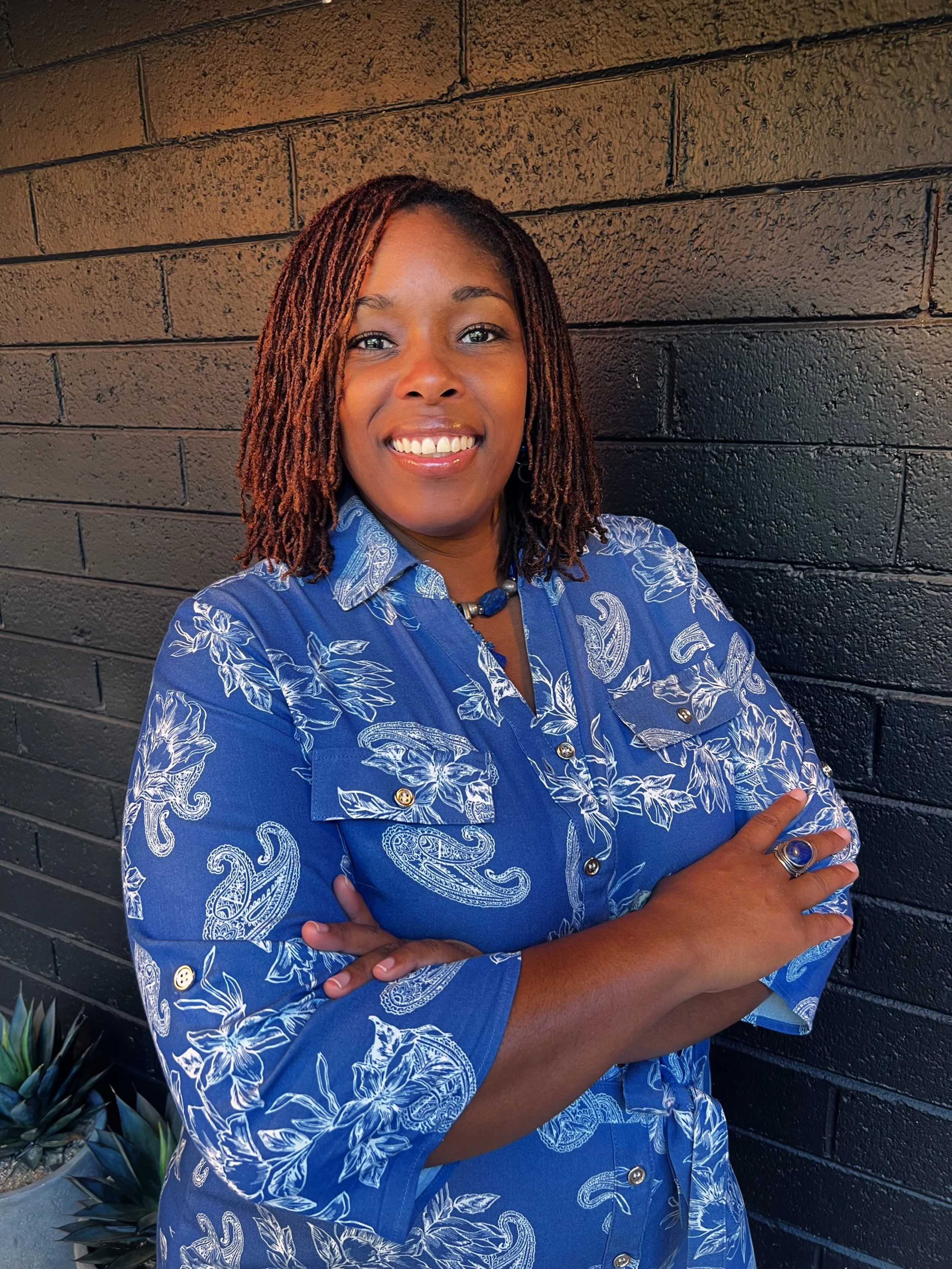 A woman with medium length curly hair wearing a blue button-up shirt with white floral and paisley patterns, standing with arms crossed against a black brick wall outdoors.