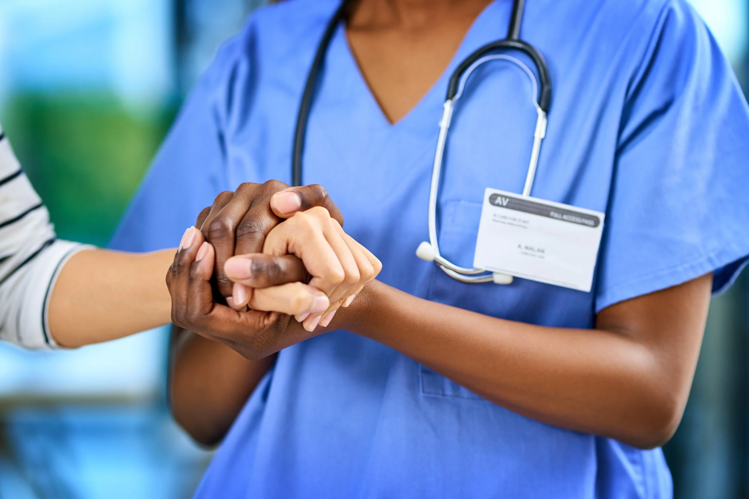 A healthcare professional in blue scrubs wearing a stethoscope, holding and shaking hands with a patient.