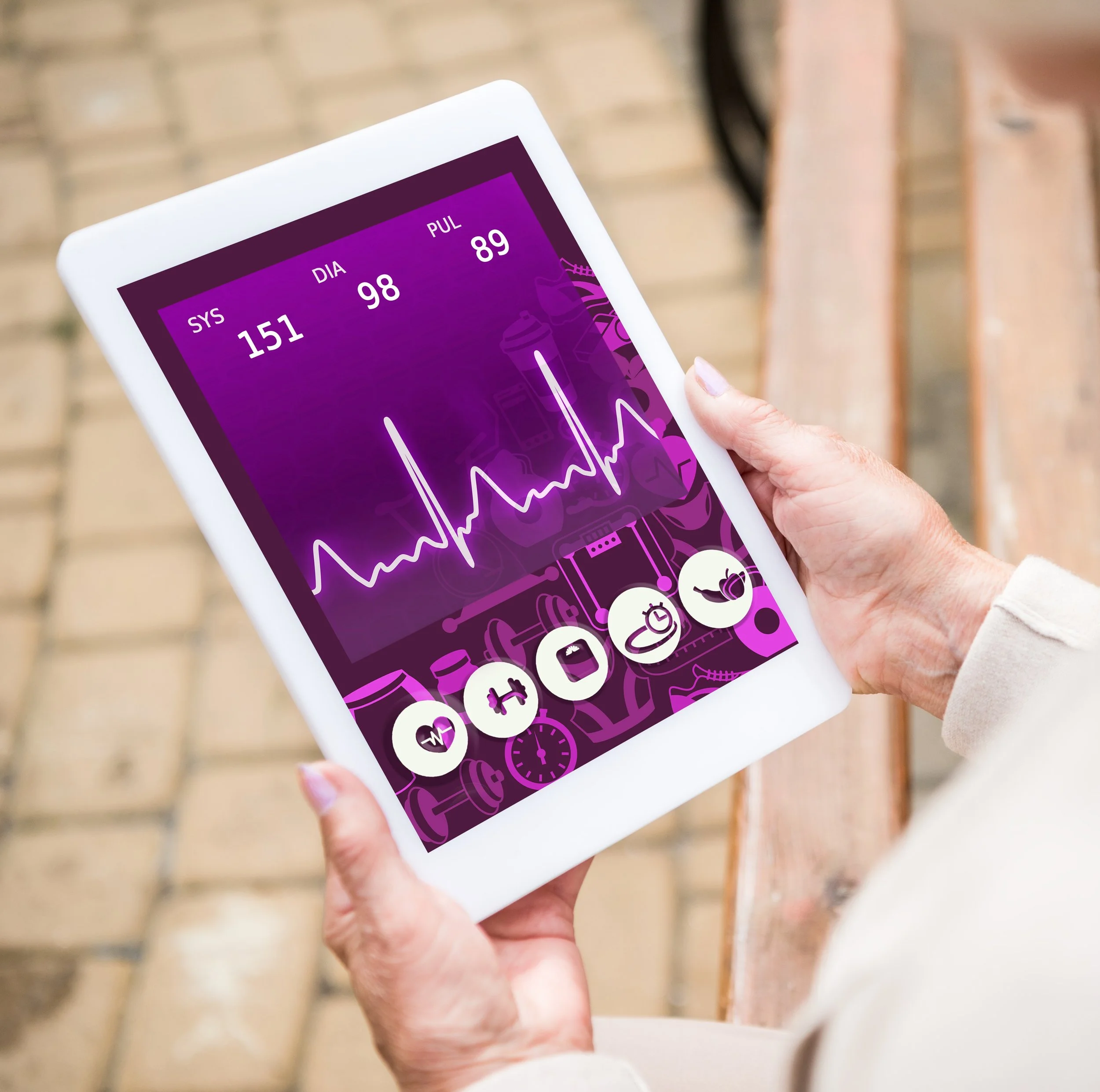 Close-up of a hand holding a white tablet device displaying health monitoring data, including blood pressure and pulse rate, against a background of a wooden surface.