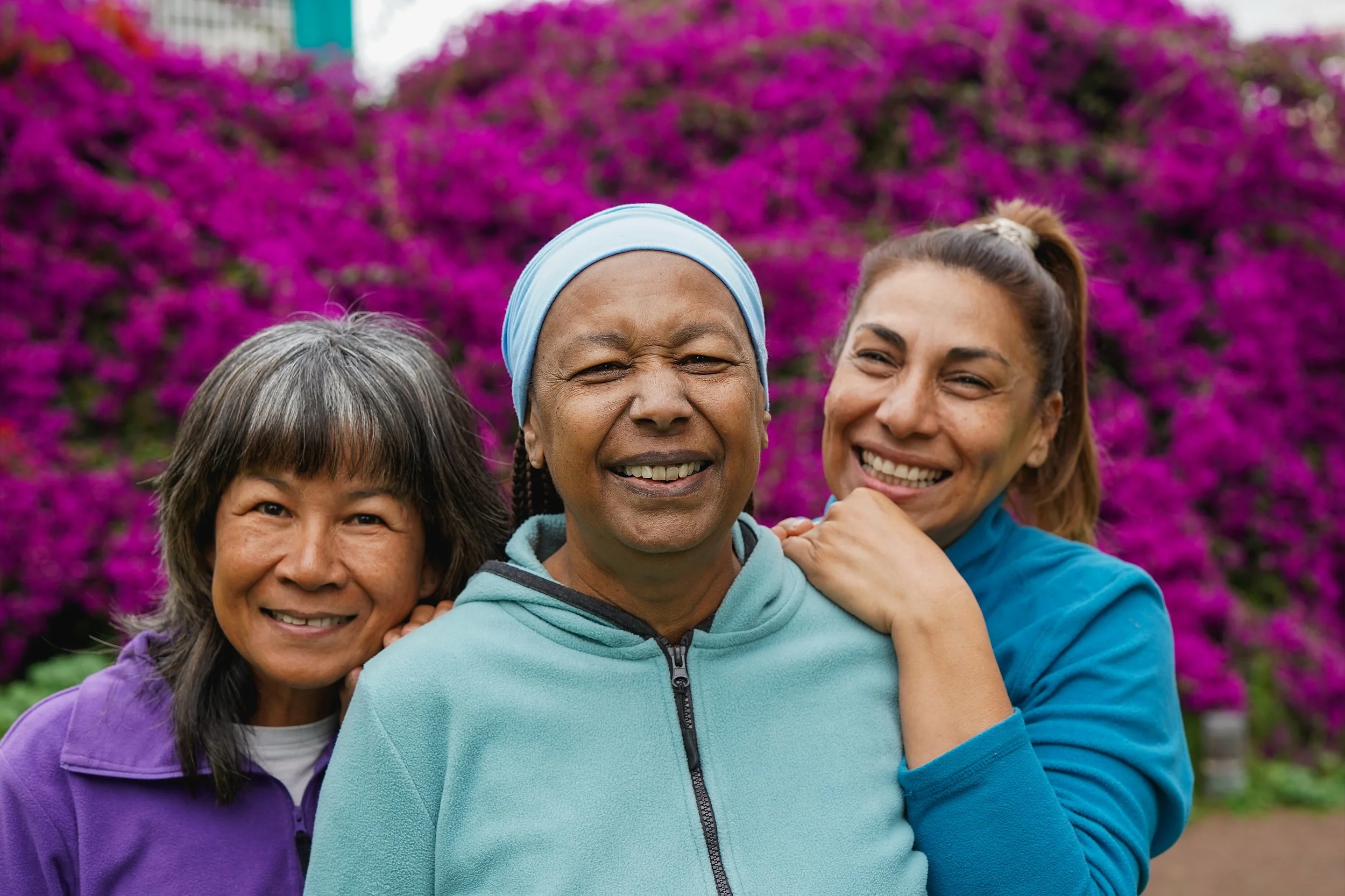 Three women standing close together outdoors with vibrant pink flowers in the background, smiling at the camera.