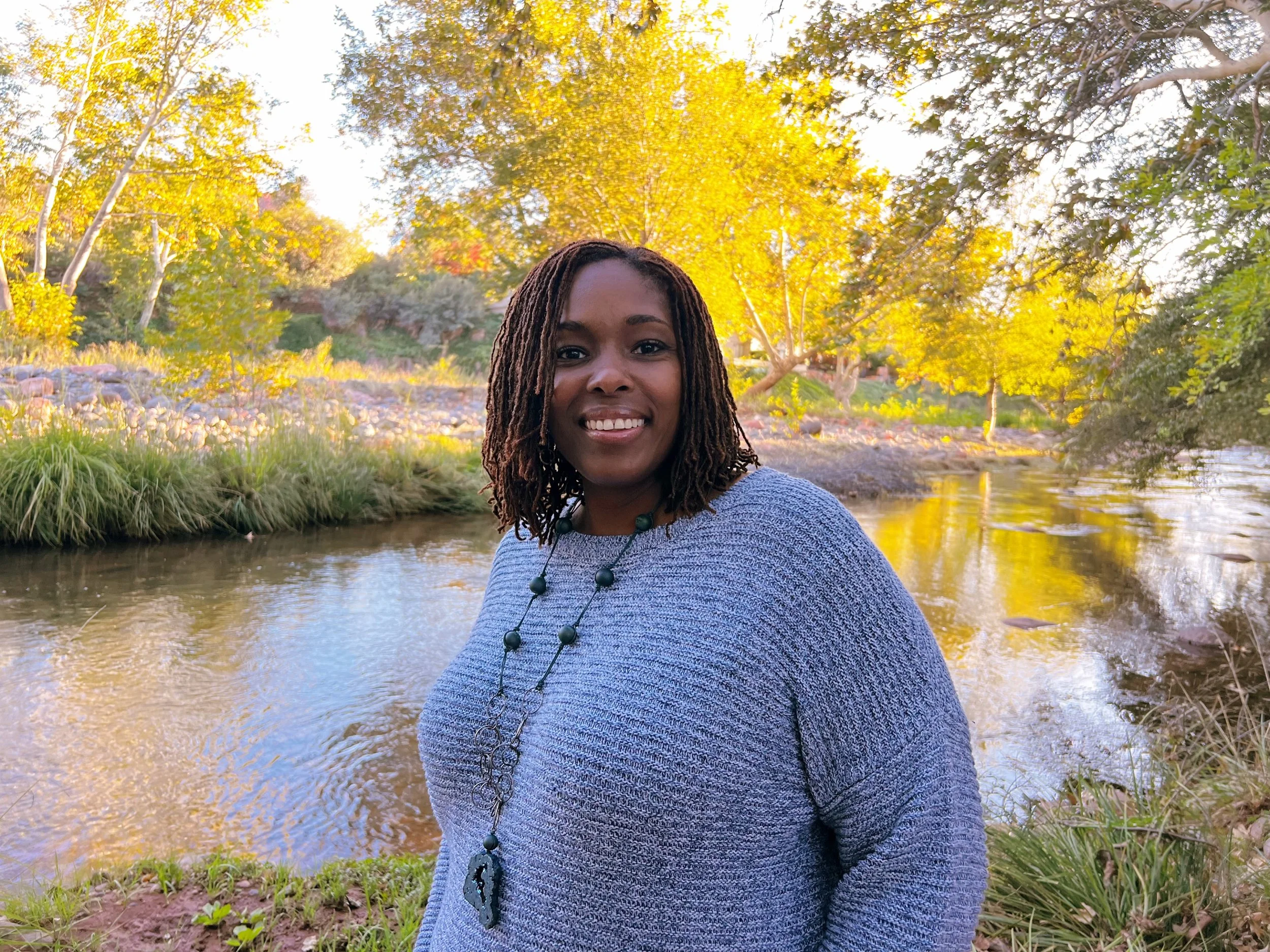 A woman with dark curly hair and a large necklace smiling outdoors near a river, with trees in fall colors in the background.
