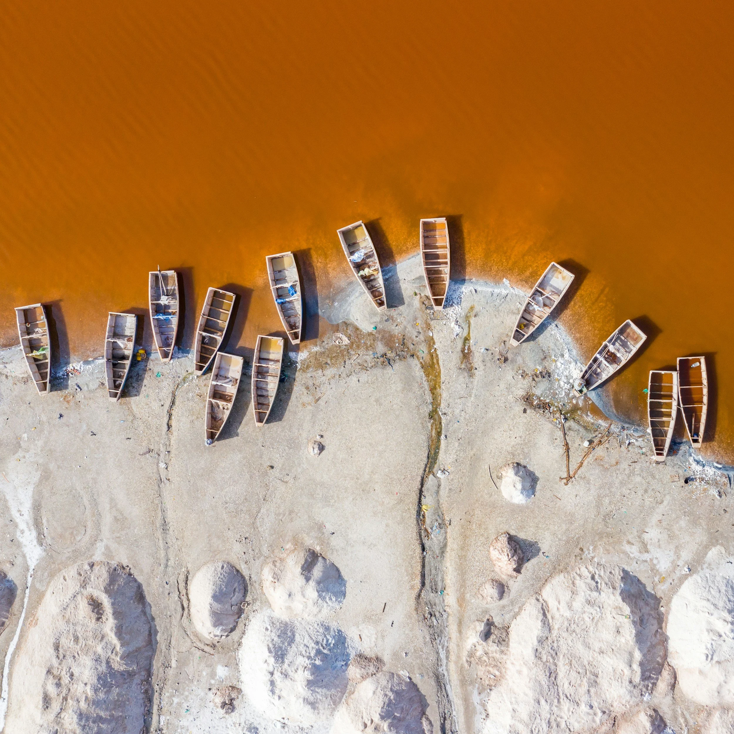 Overhead aerial view of a shoreline with several brown boats lined up along the shore, which has sandy and rocky terrain, with the water appearing reddish-brown.