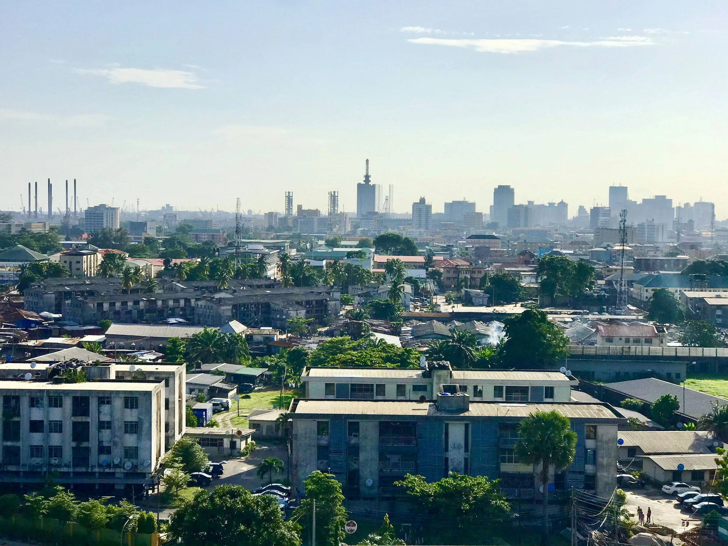 A cityscape with mid-rise buildings, trees, and a distant skyline, under a clear sky with some clouds.