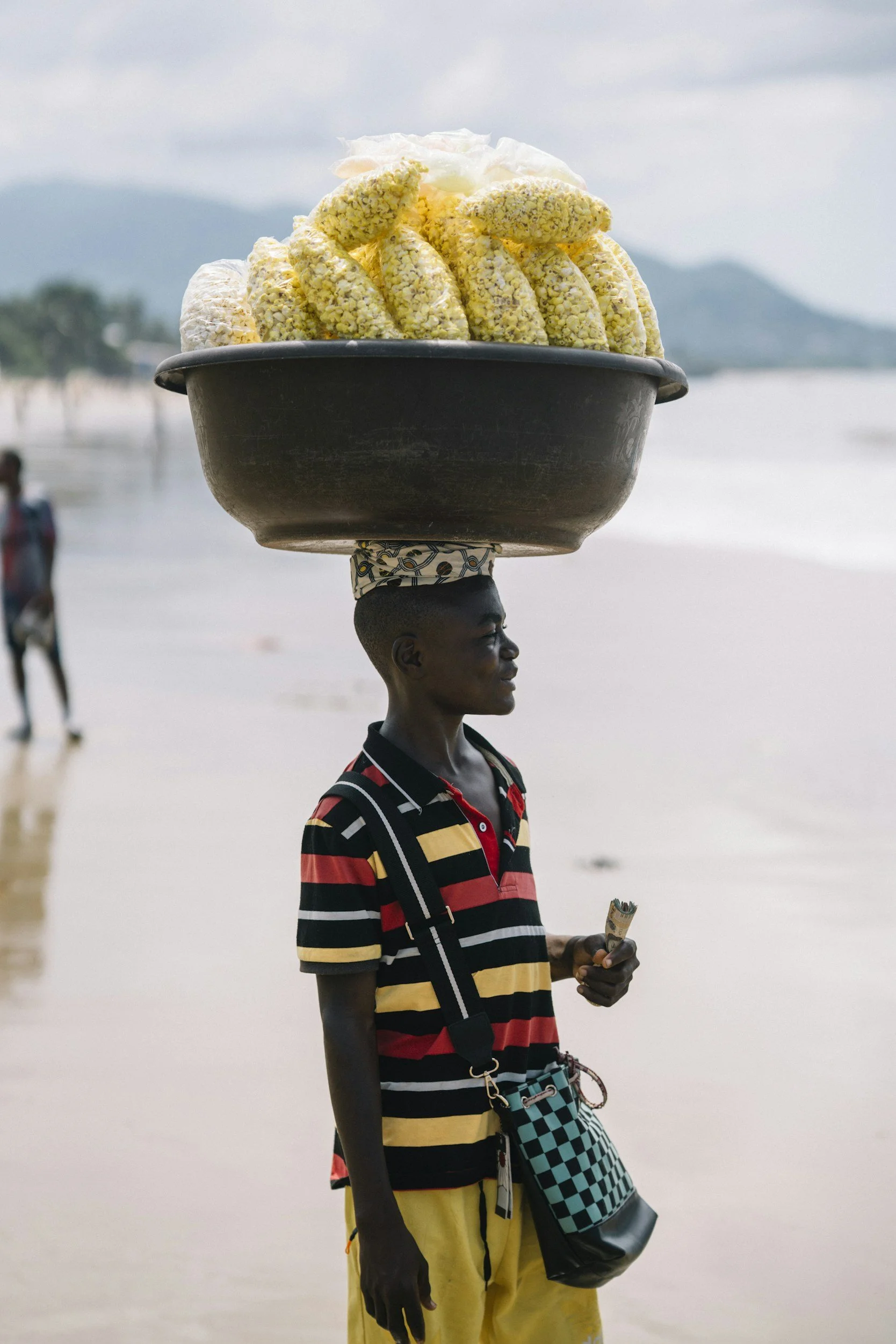 A young man with dark skin balancing a large black bowl filled with bananas and other items on his head, standing on a beach with an ocean and mountains in the background, wearing a striped shirt and yellow pants.