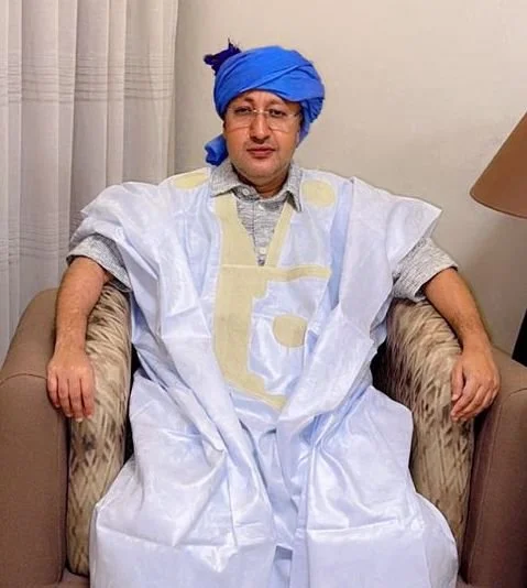 A man dressed in traditional Indian attire, wearing a blue turban, sitting on a beige armchair with a calm expression.