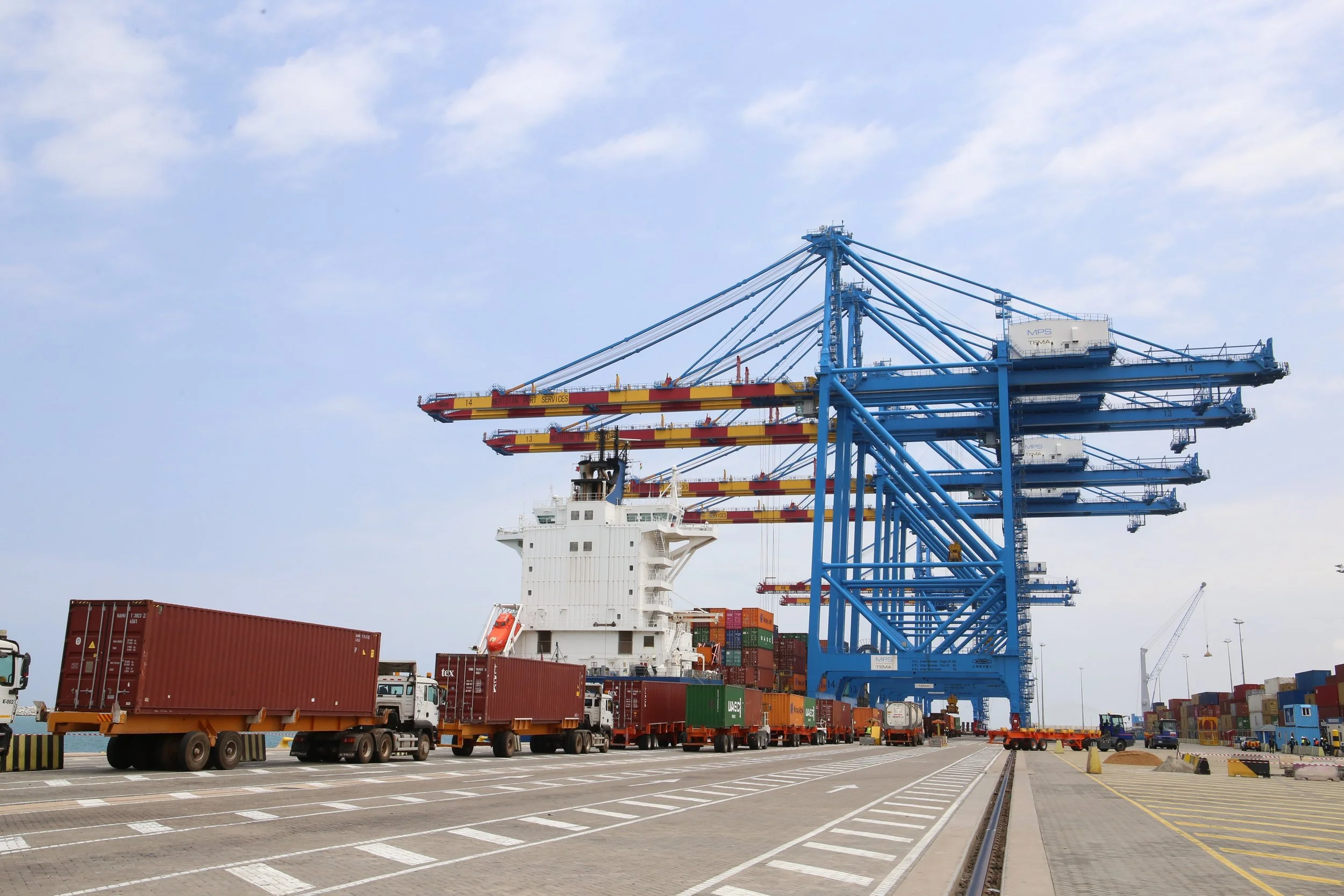 Cargo port with blue gantry cranes, trucks, and shipping containers under a partly cloudy sky.