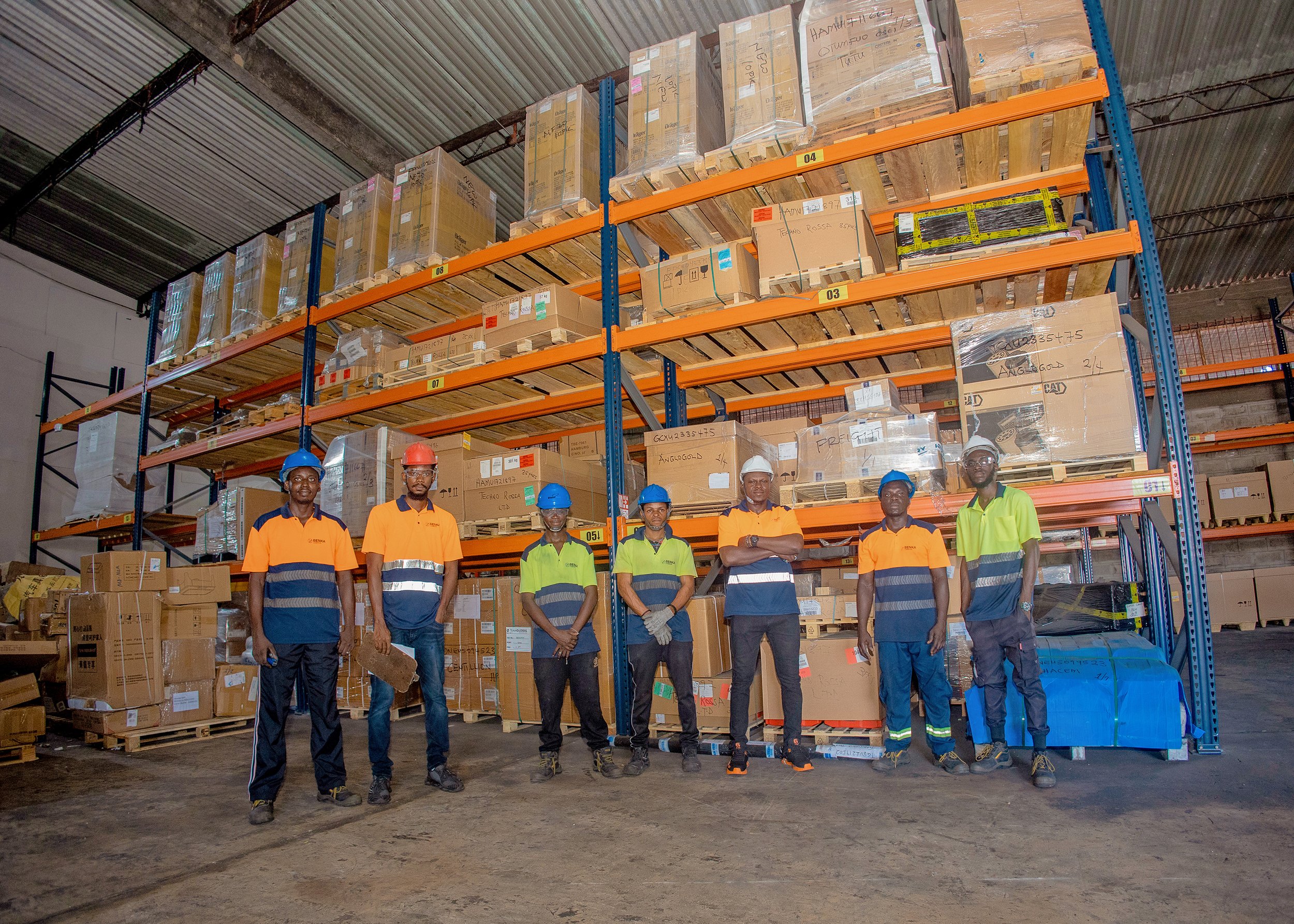 Six workers in high-visibility orange and yellow shirts and safety helmets standing inside a warehouse in front of orange and blue metal shelves filled with boxes and pallets.