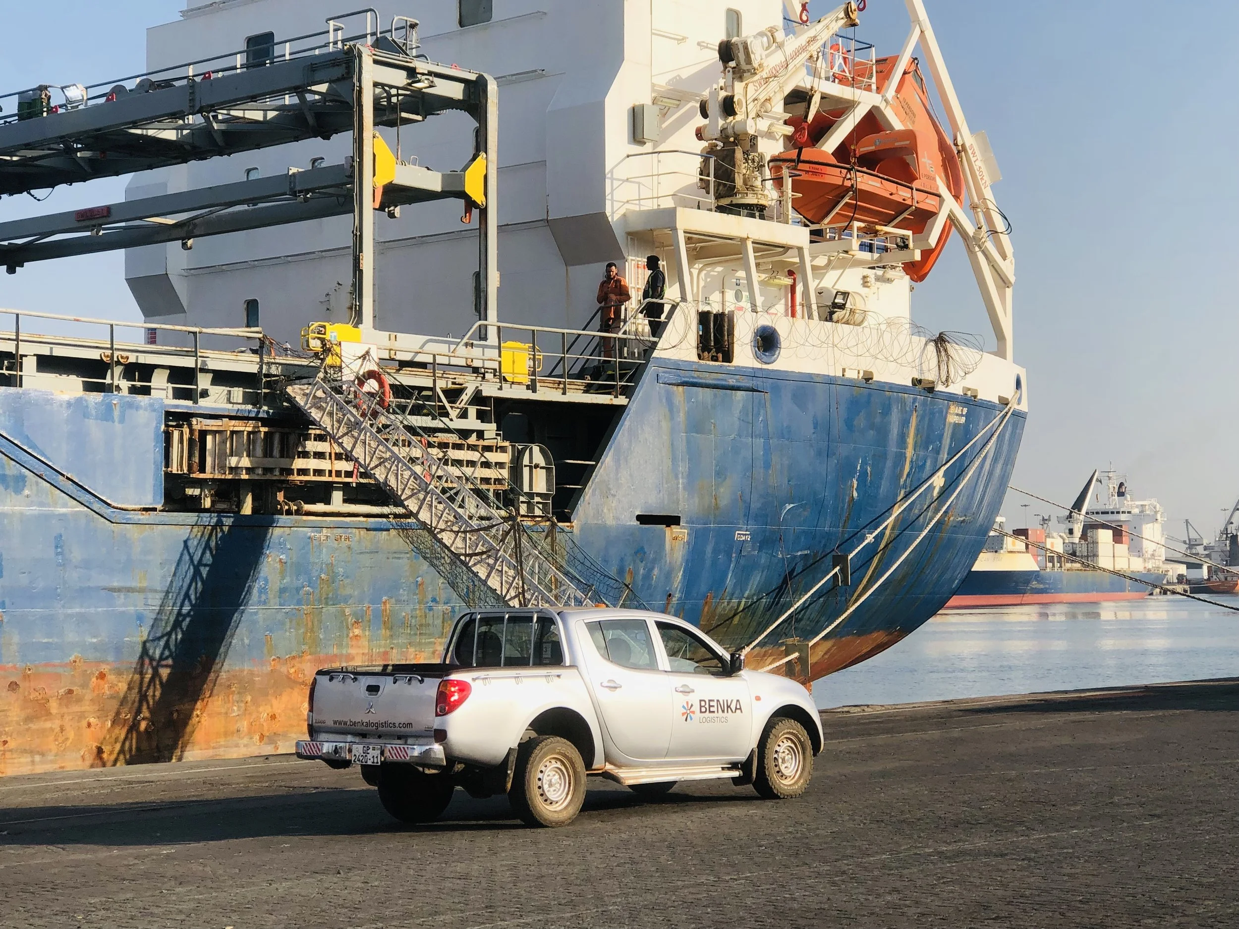 A large cargo ship docked at a port with a white truck labeled Benka Logistics parked nearby. The ship has a blue hull and various equipment and life rafts on deck, with a ladder leading from the ground to the ship's side.