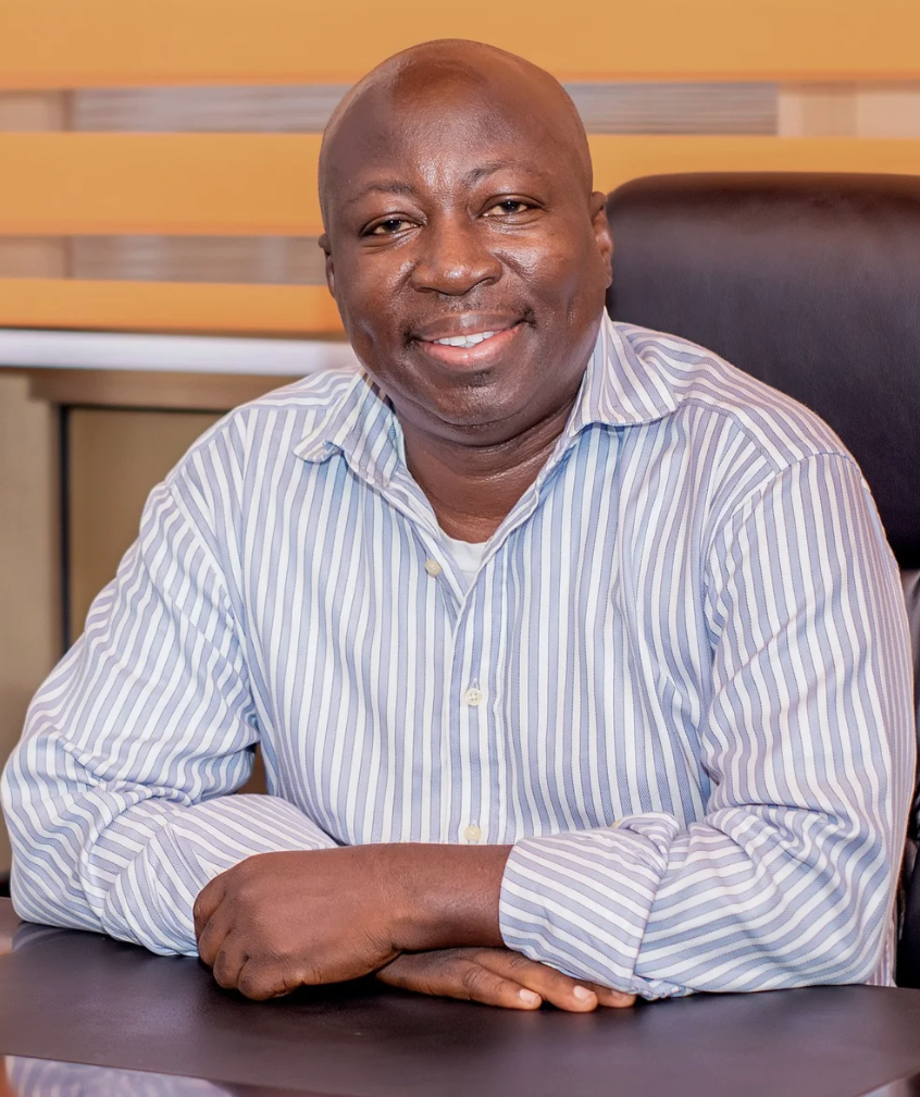 A smiling man sitting at a desk in an office, wearing a light blue and white striped shirt.