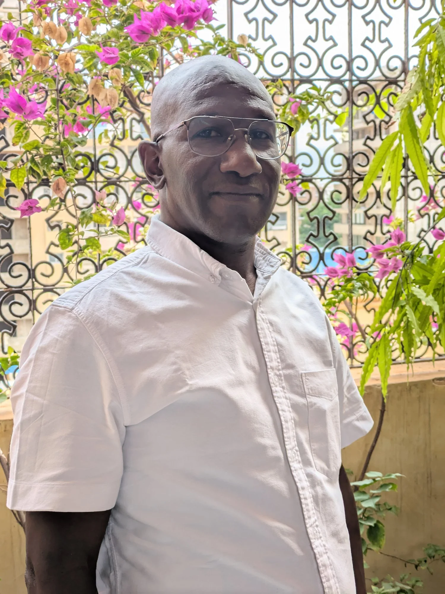 A man wearing glasses and a white short-sleeved shirt standing on a balcony with pink flowers and decorative wrought iron railing in the background.