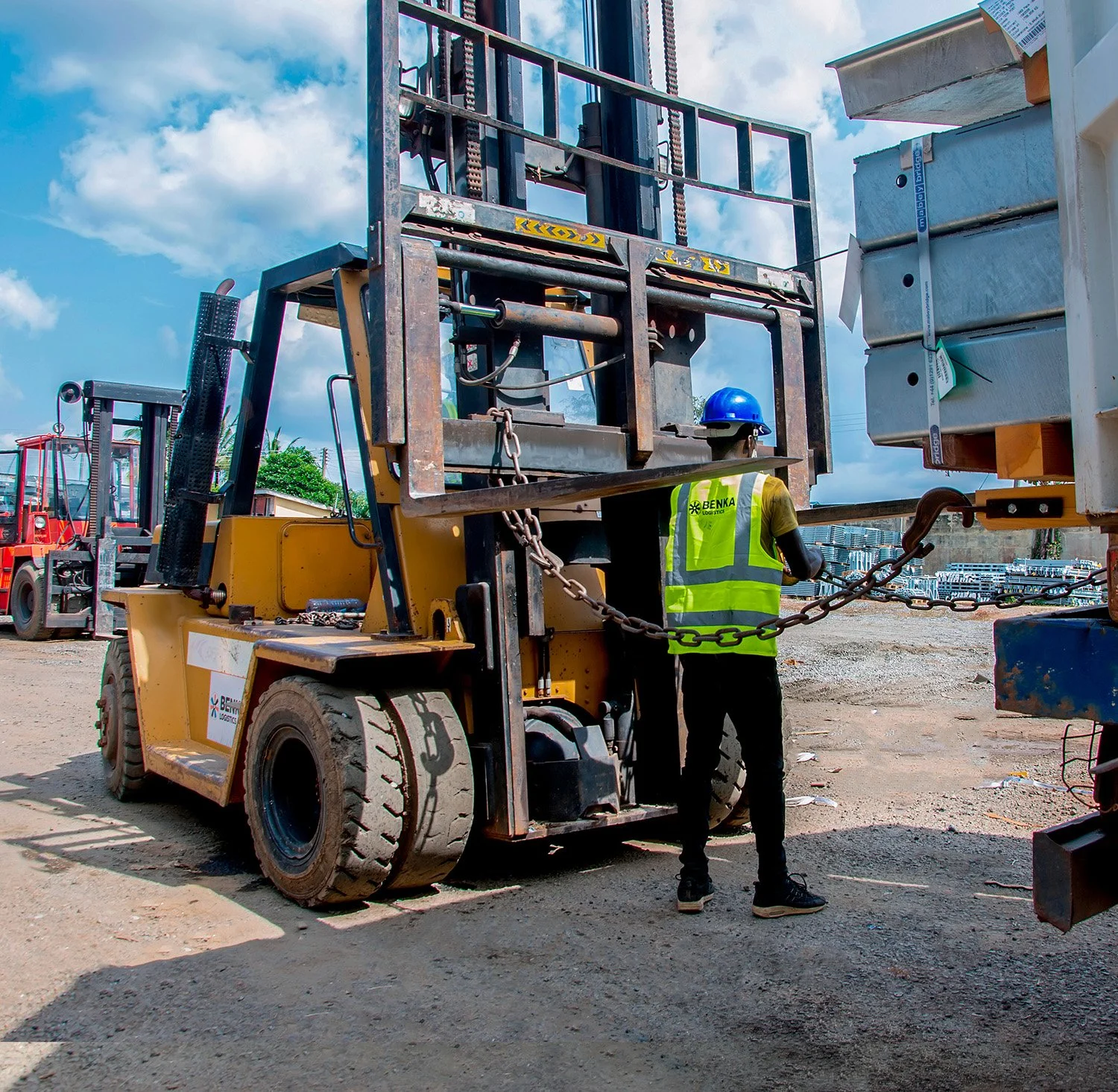 Construction worker in safety vest and helmet checks a forklift at a construction site with blue sky and clouds in the background.