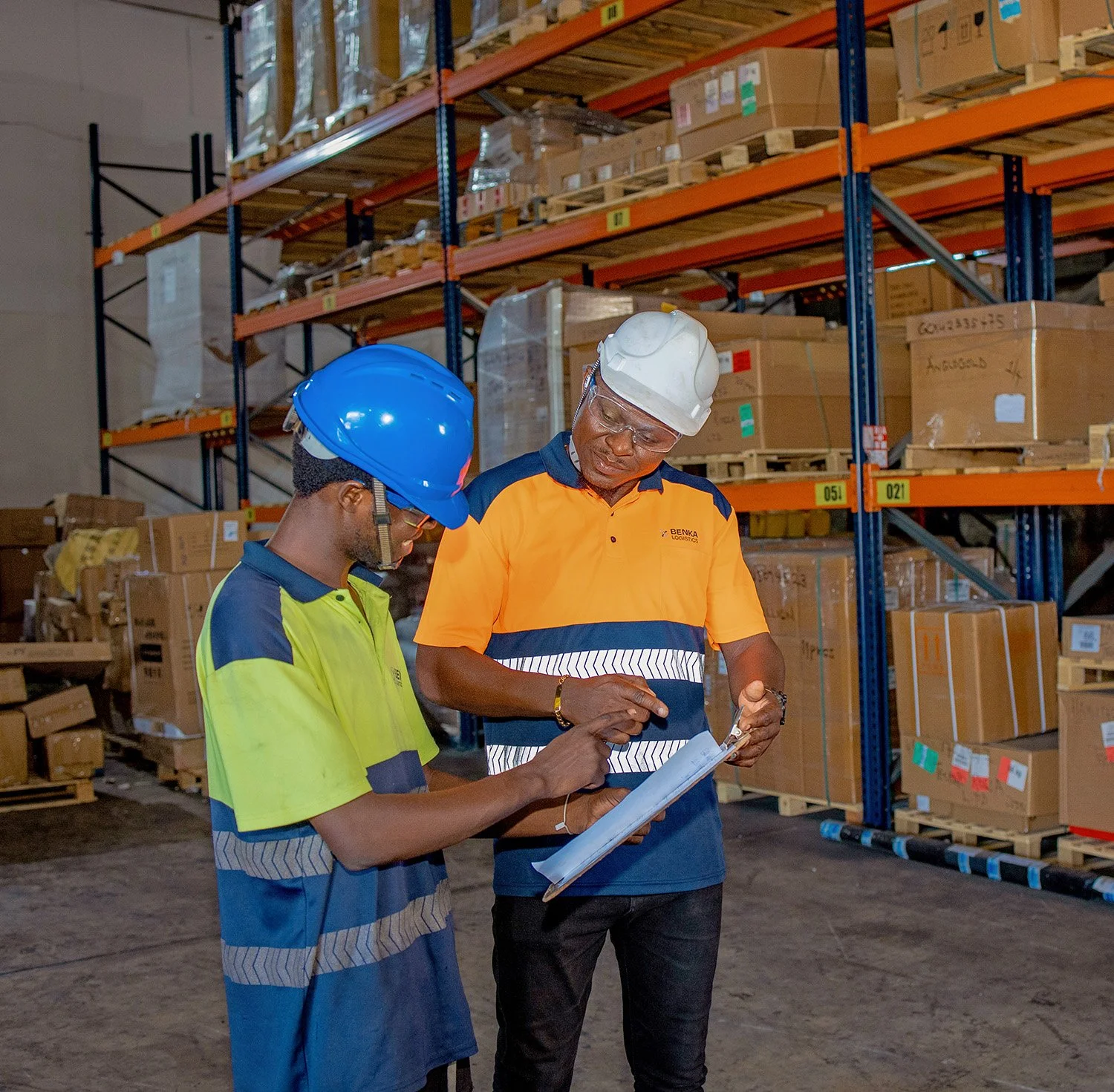 Two workers wearing safety helmets and high-visibility clothing, standing in a warehouse aisle, reviewing documents on a clipboard.