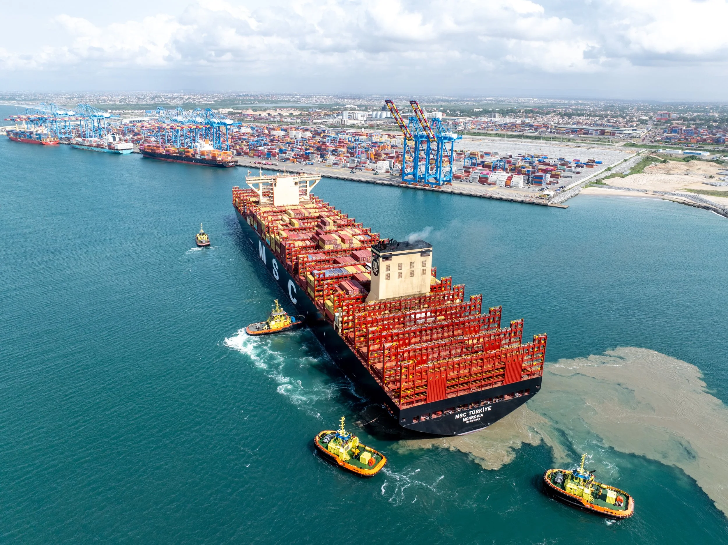 Aerial view of a large container cargo ship traveling through a harbor, with tugboats guiding it. The harbor has numerous warehouse cranes and stacks of shipping containers, with a cityscape and distant mountains in the background.