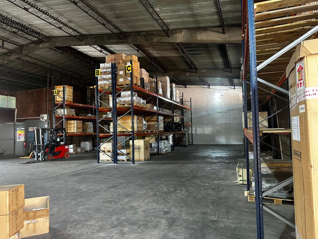 Inside a warehouse with metal shelving holding stacked boxes and pallets, a red forklift parked on the left, and a large empty space in the center.