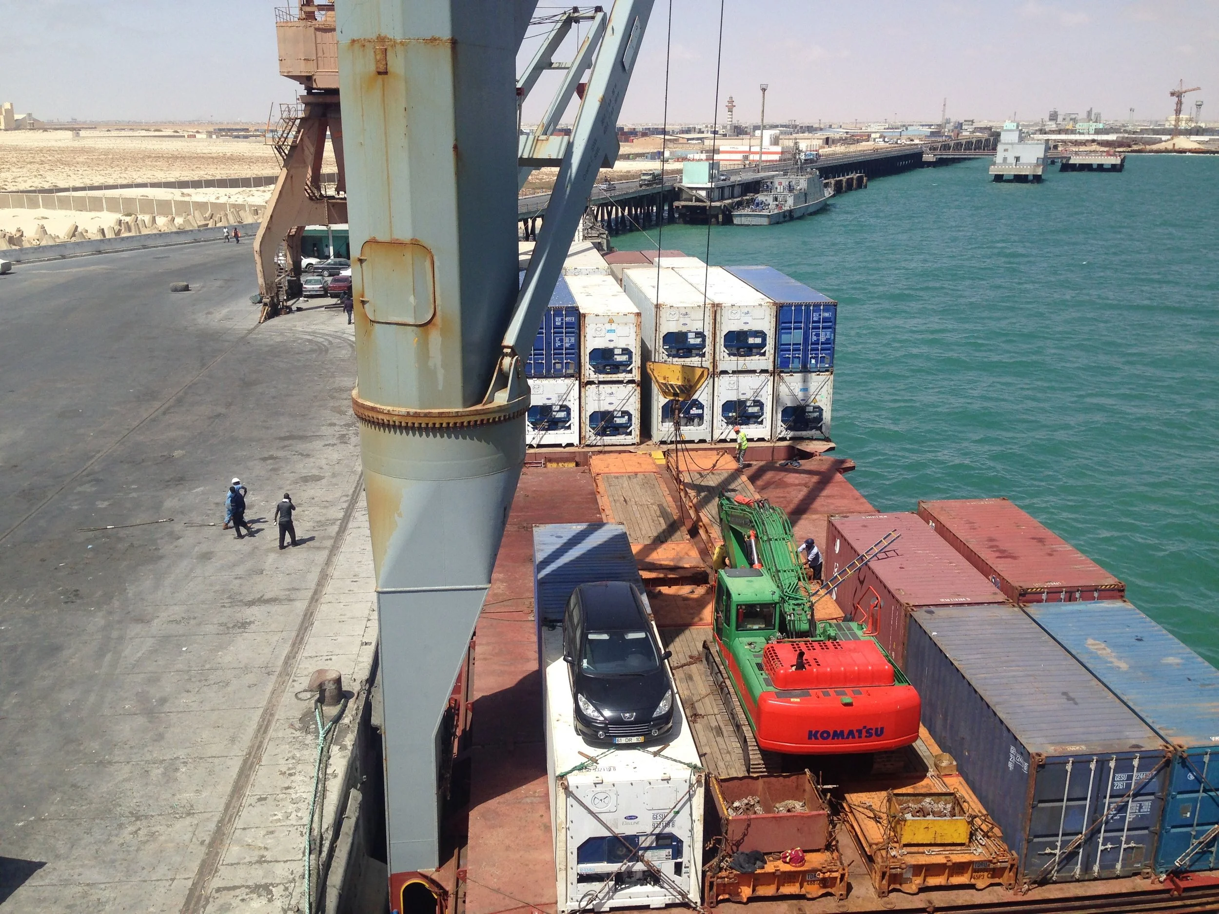 Cargo port with containers, cranes, a small green and red crane, trucks, cars, and workers near the water, with a sandy desert and distant buildings in the background.