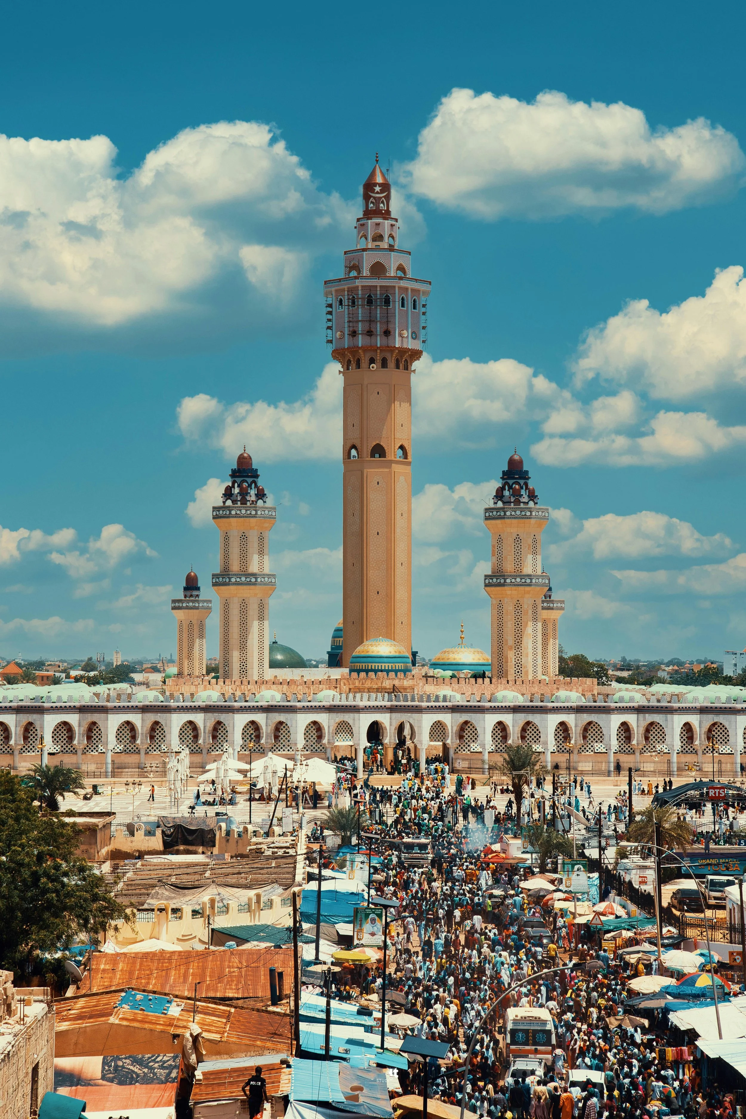 A large mosque with multiple minarets under a partly cloudy sky, with a crowded marketplace in the foreground.