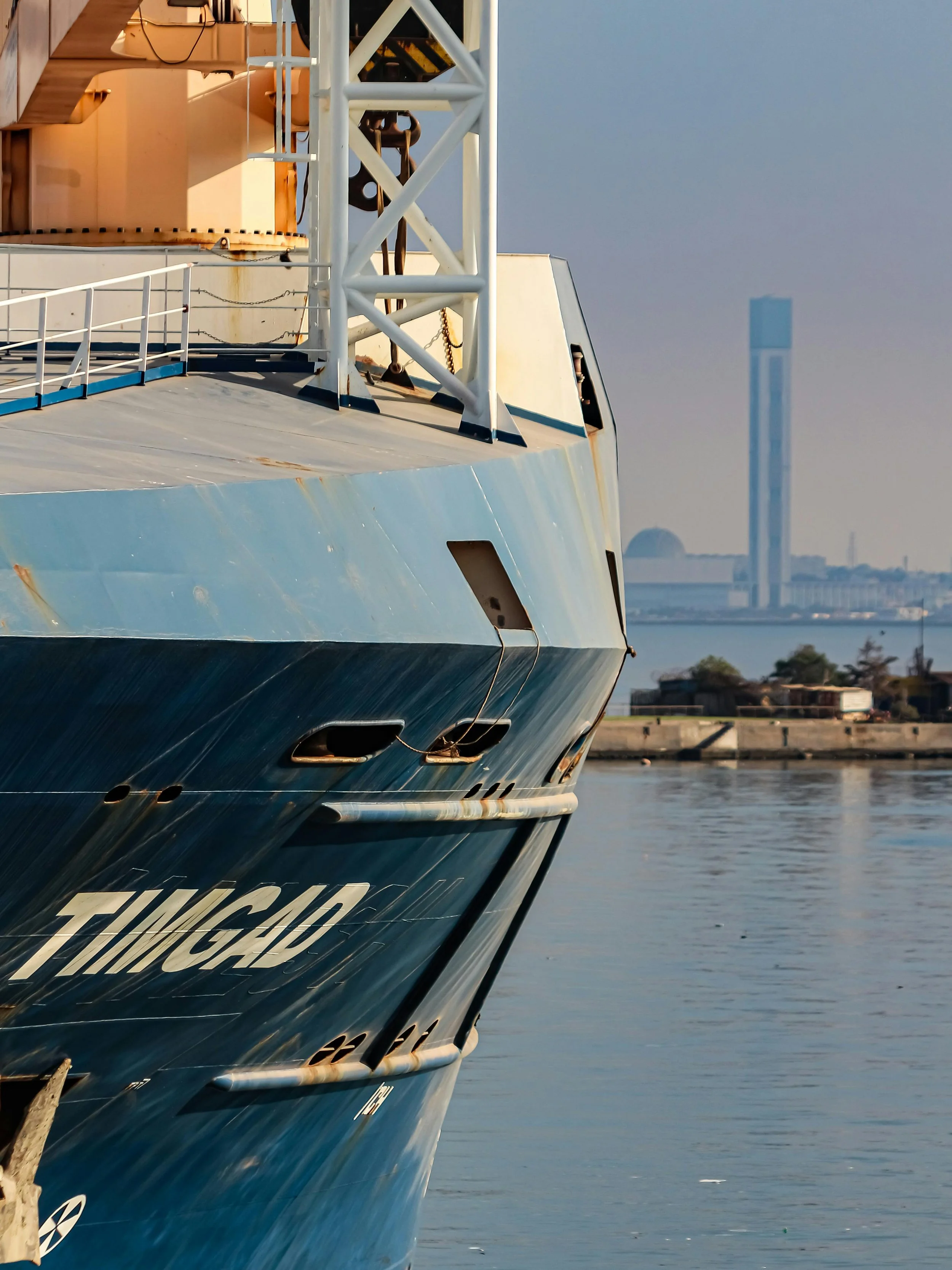 Close-up of the bow of a large blue ship docked at a harbor, with some water and city skyline in the background.