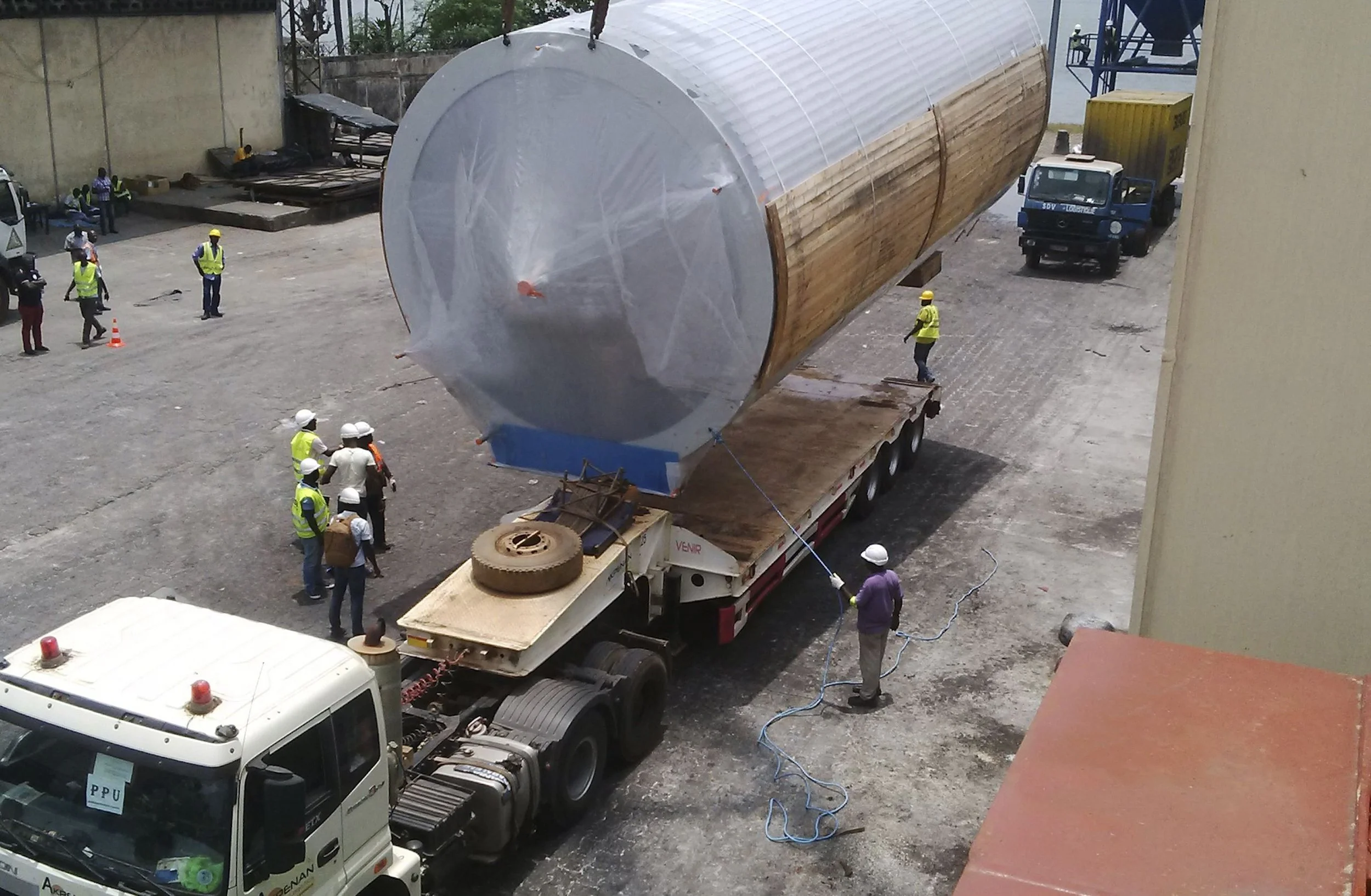 A large cylindrical tank being transported on a flatbed trailer, surrounded by workers in safety gear at an industrial site.