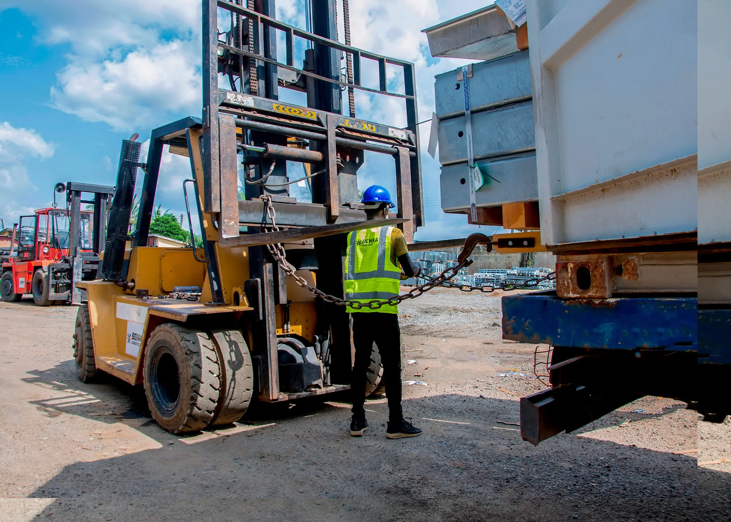 Construction worker in a high-visibility vest and blue helmet operating a forklift at a construction site with other forklifts in the background.