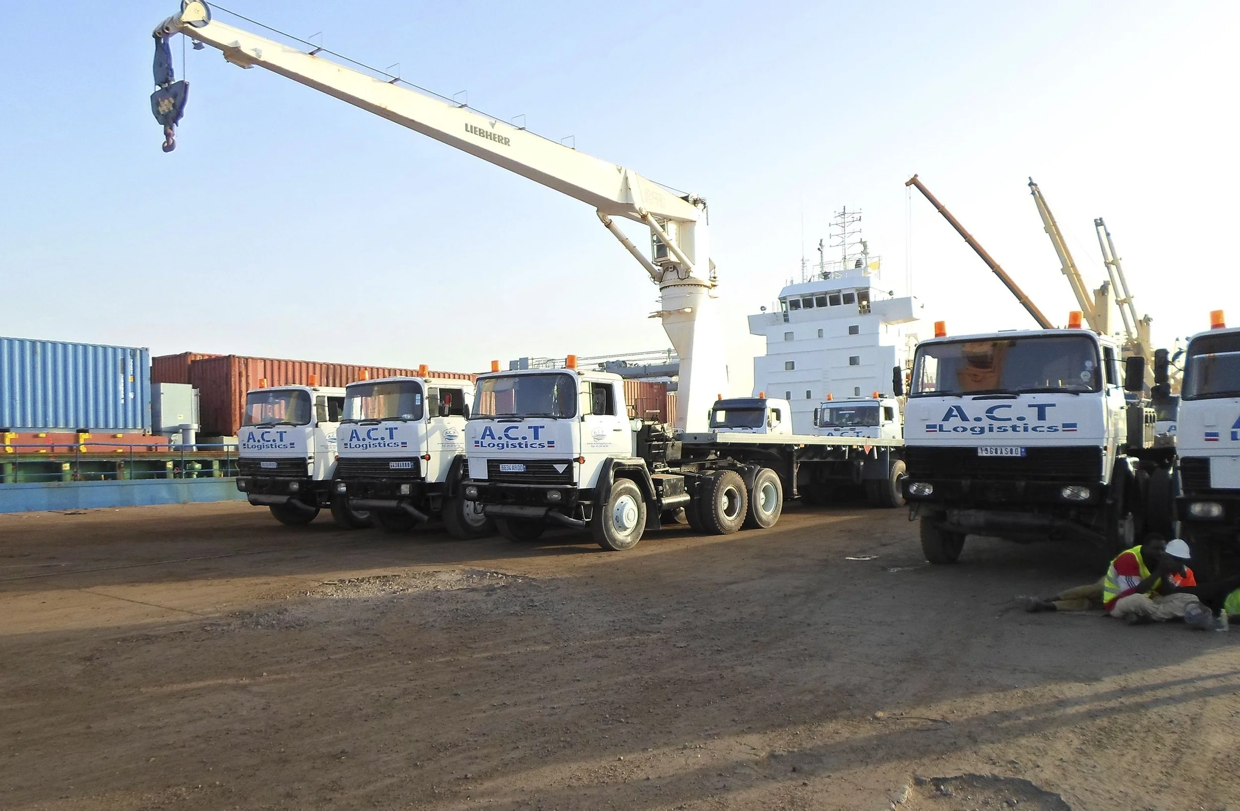 Construction site with trucks labeled A.C.T Logistics, containers, cranes, and workers wearing safety gear.