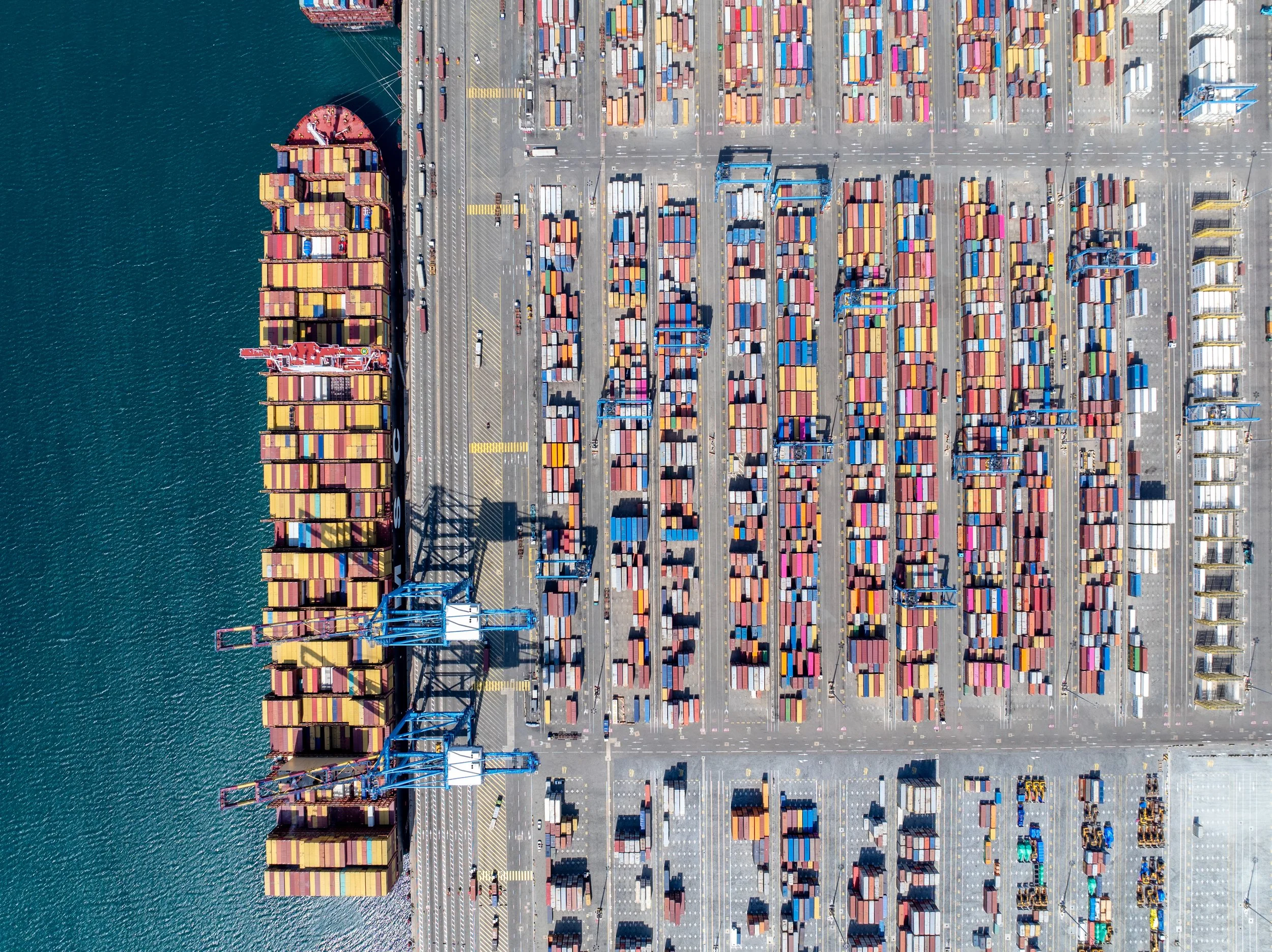 Aerial view of a busy container port with colorful cargo containers, cranes, and ships by the water.