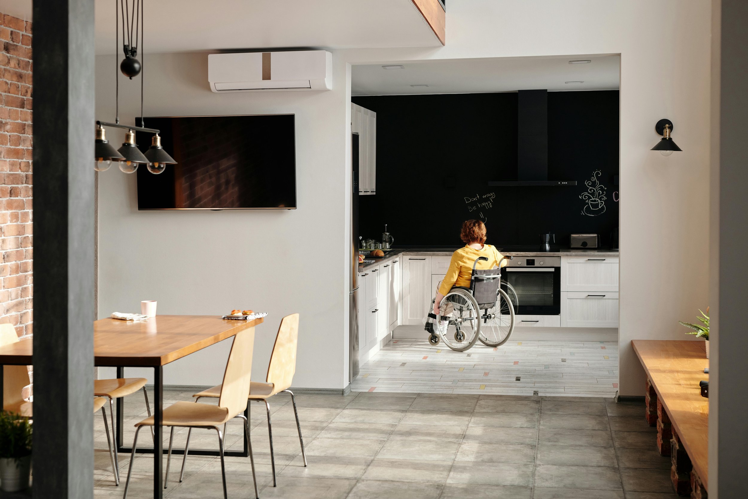 A woman in a yellow shirt sitting in a wheelchair in a modern kitchen area with white cabinets, a black chalkboard wall, and a black vent hood. The space is bright with natural light, and there is a dining area with a wooden table and beige chairs on the left.